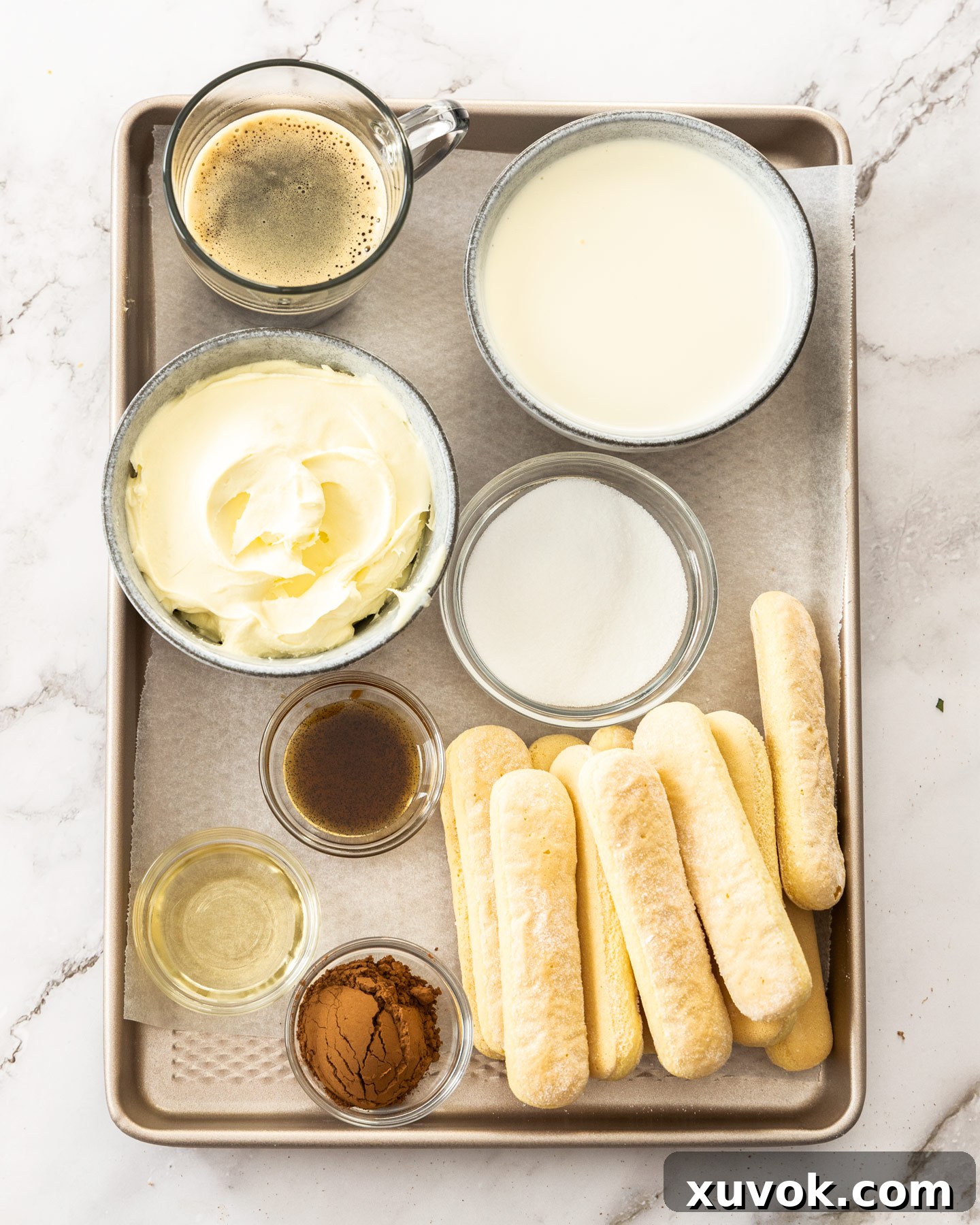 All the essential ingredients for making tiramisu cups, neatly arranged on a baking tray. This includes cold heavy cream, mascarpone cheese, caster sugar, vanilla extract, ladyfingers, freshly brewed espresso, a bottle of Frangelico liqueur, and cocoa powder.