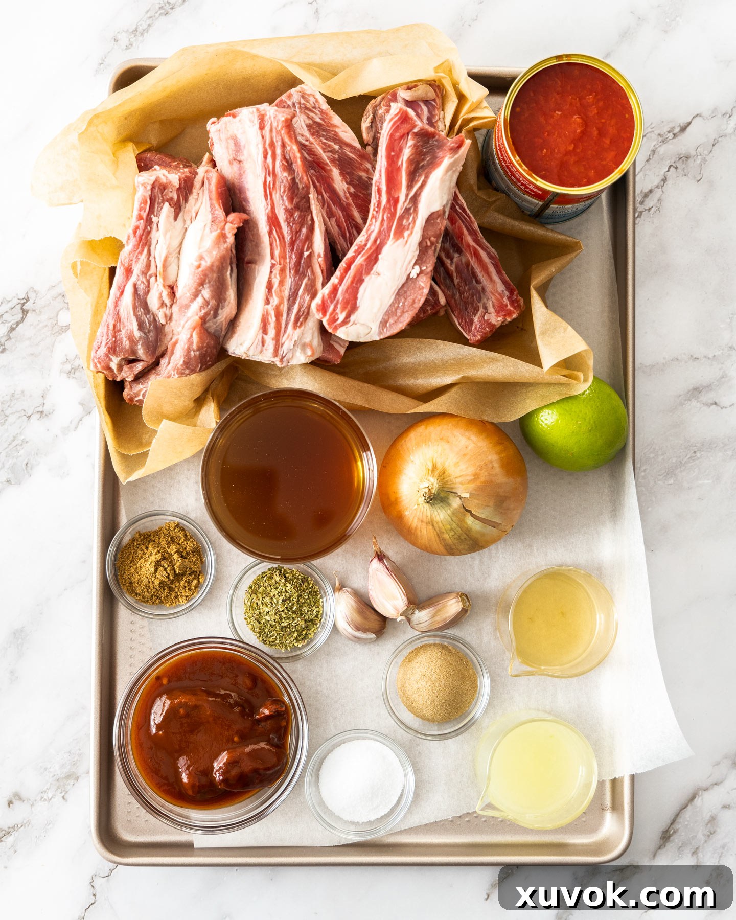 A selection of fresh ingredients for Mexican short ribs laid out on a baking tray, including beef, tomatoes, and spices.