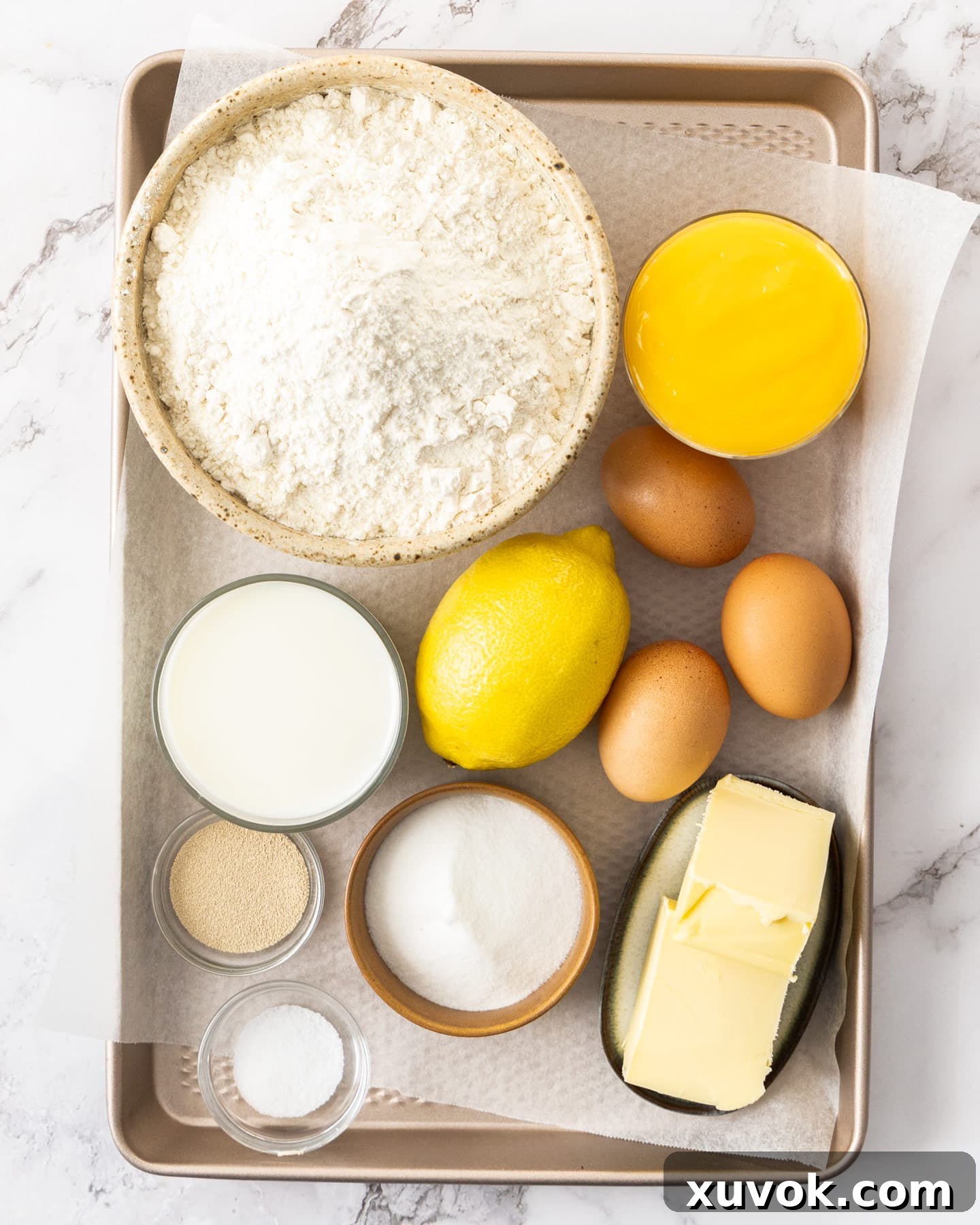 Fresh ingredients for making lemon donuts, including milk, sugar, yeast, flour, eggs, butter, lemons, and lemon curd, arranged neatly on a metal tray.