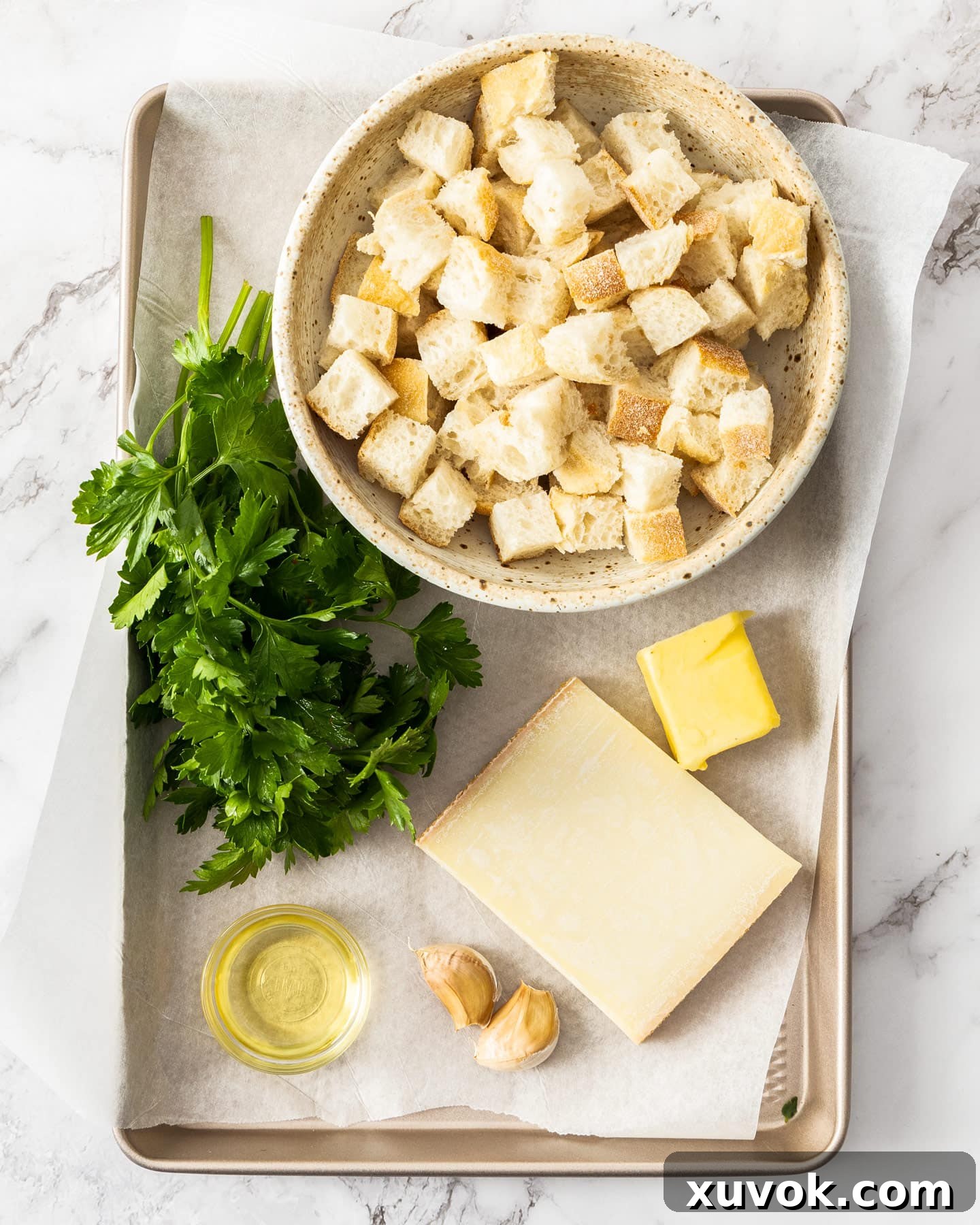 Ingredients for cheese garlic croutons laid out on a baking tray: bread cubes, garlic, butter, olive oil, Gruyère cheese, and parsley.