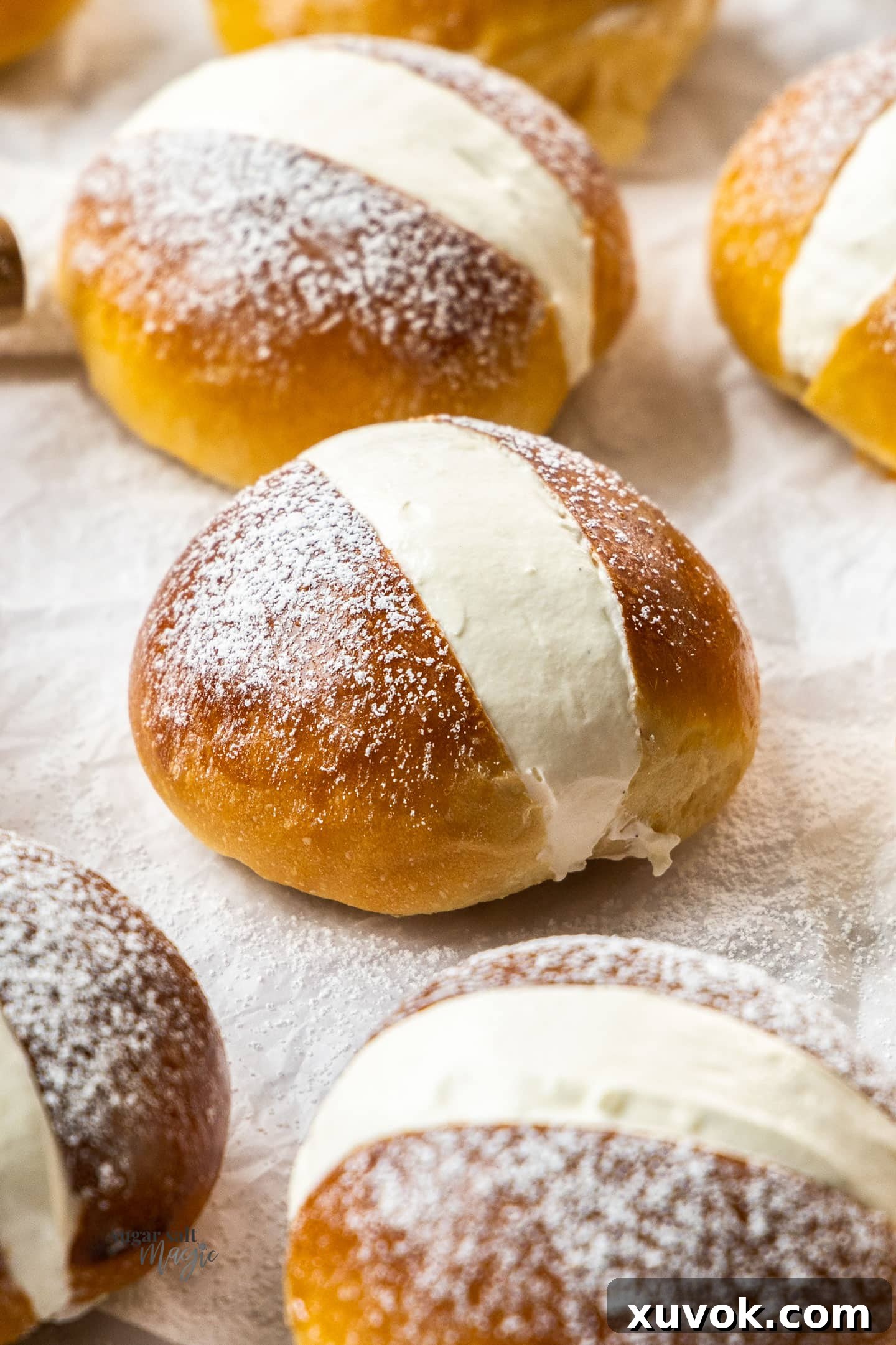 A close-up view of a freshly filled maritozzo bun, with more cream buns softly blurred in the background, showcasing their soft texture and generous cream filling. The image evokes a sense of delectable indulgence.