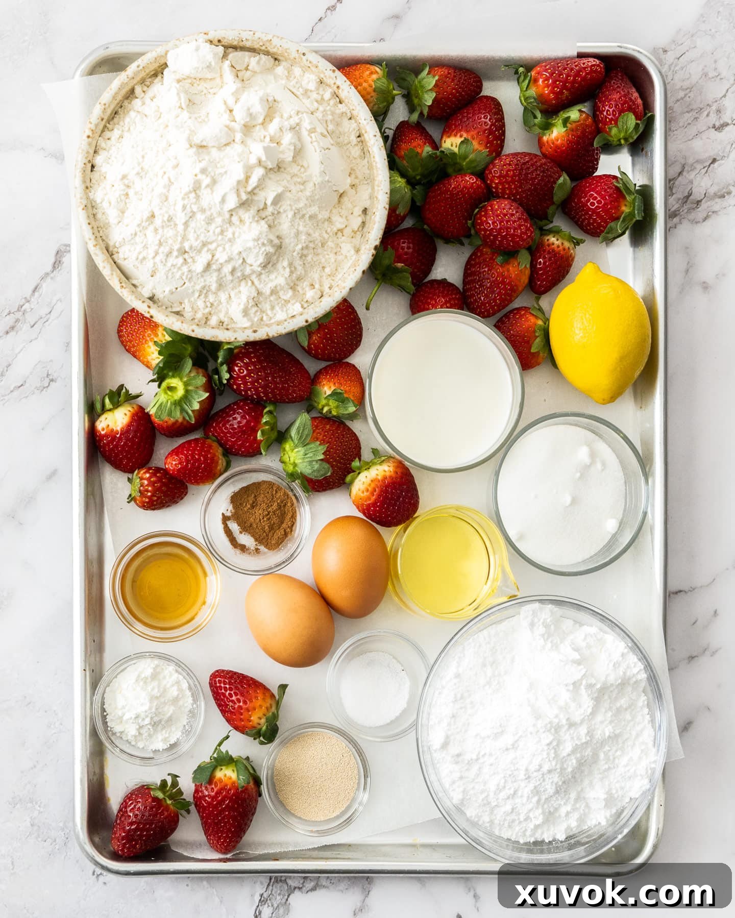 A collection of fresh ingredients for strawberry cinnamon rolls, including flour, milk, eggs, vanilla, and a bowl of vibrant strawberries, arranged on a baking tray.