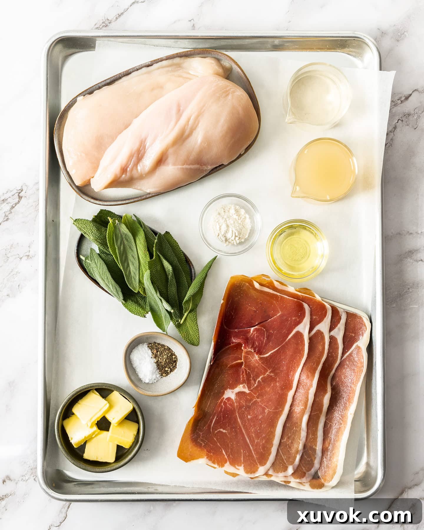 A tray filled with all the ingredients required for chicken saltimbocca, including chicken breasts, prosciutto, sage, butter, flour, wine, and stock.