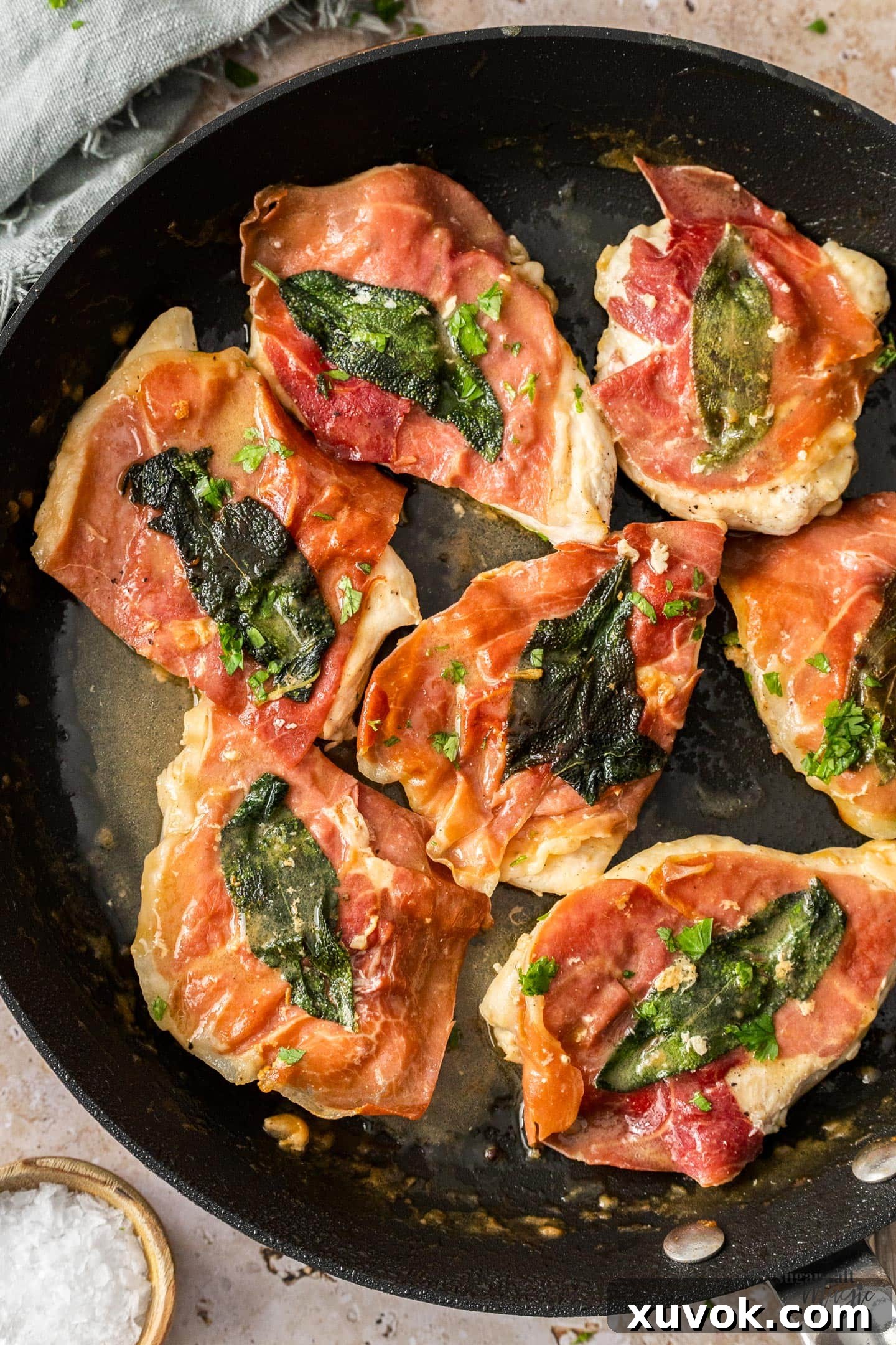 Seven perfectly cooked pieces of chicken saltimbocca, topped with crispy prosciutto and sage, nestled in a rich pan sauce within a skillet.