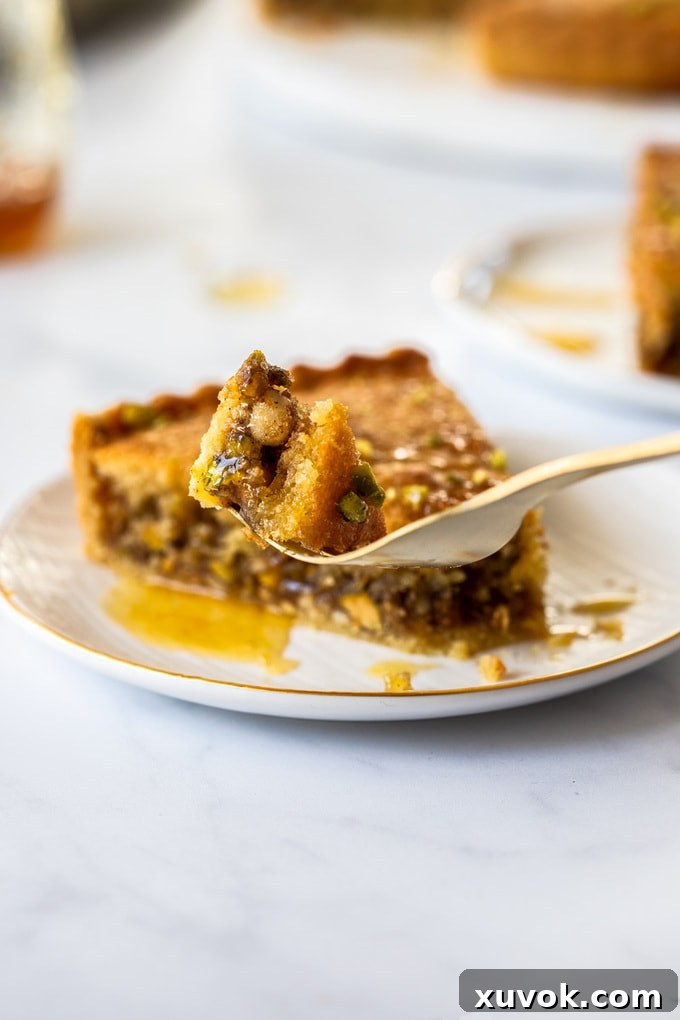 A close-up shot of a forkful of Honey Baklava Tart, showing the layers of the tender frangipane, nuts, and crisp crust, with the rest of the slice on a white plate in the background.