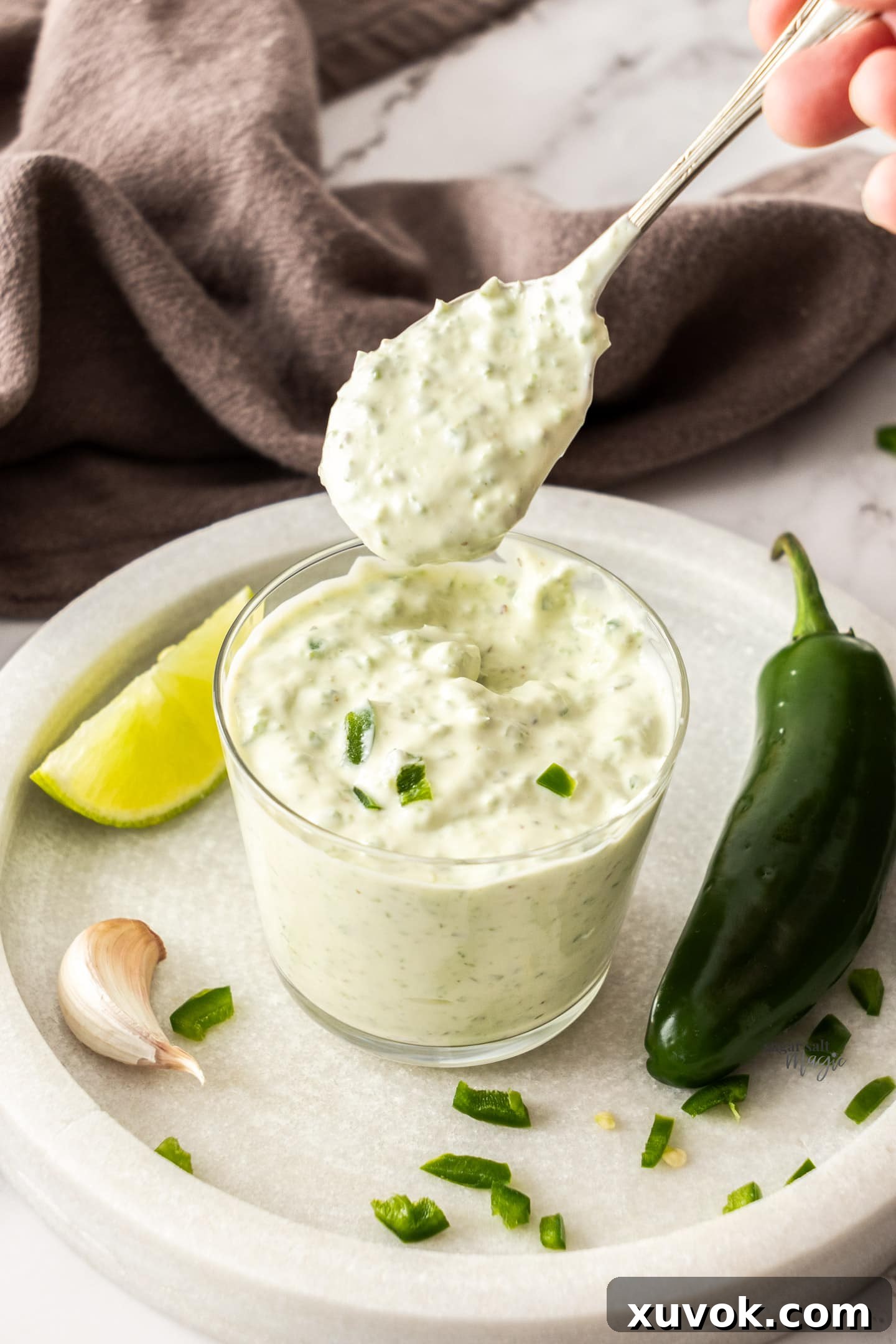 A spoon taking a scoop of creamy jalapeno mayo out of a clear glass jar.