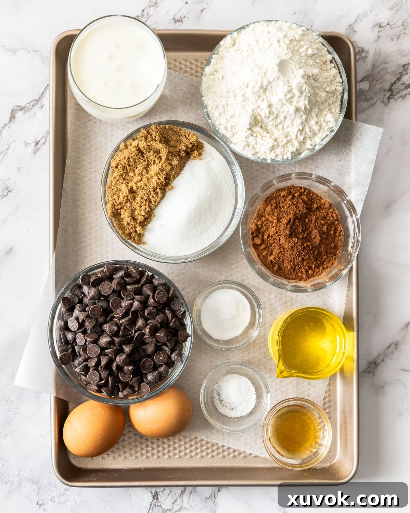 A flat lay photograph of all the essential ingredients for making triple chocolate muffins, neatly arranged on a rustic baking tray, including flour, cocoa powder, sugars, chocolate chips, eggs, buttermilk, oil, and vanilla extract.