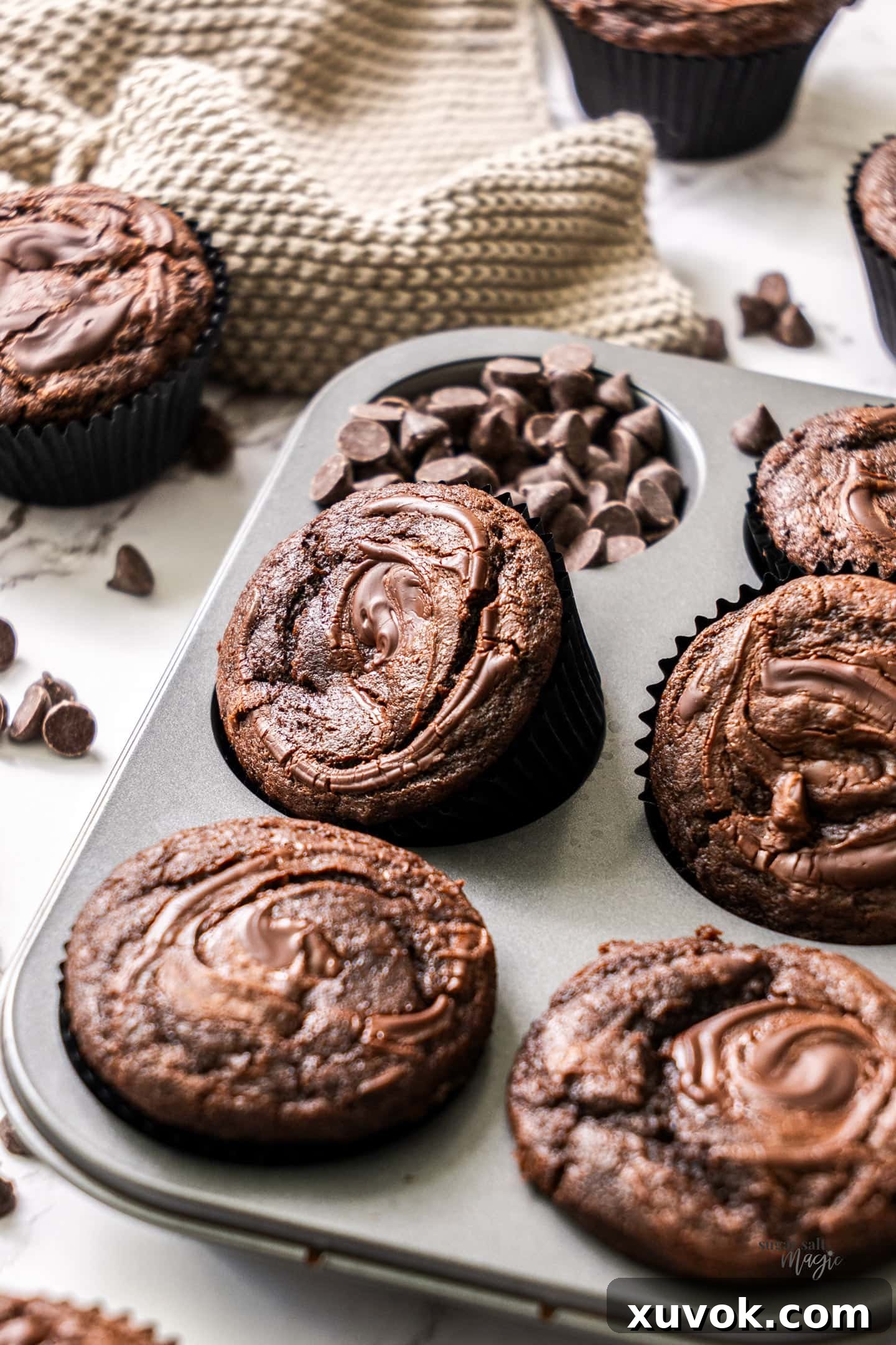 A single triple chocolate muffin, perfectly domed with a cracked top and a glistening chocolate drizzle, resting at an angle in a muffin pan, highlighting its appealing texture.
