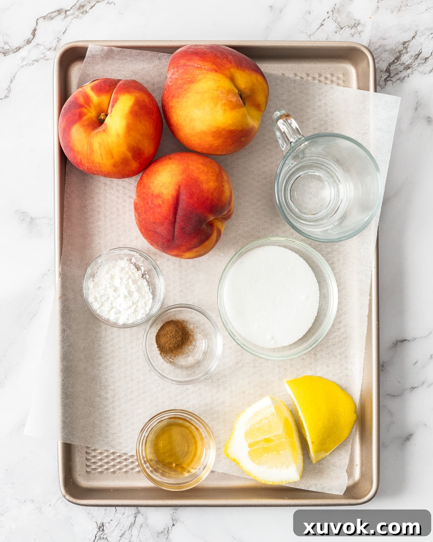 Fresh ingredients for peach compote, including ripe peaches, sugar, lemon, vanilla, and cinnamon, neatly arranged on a baking tray.