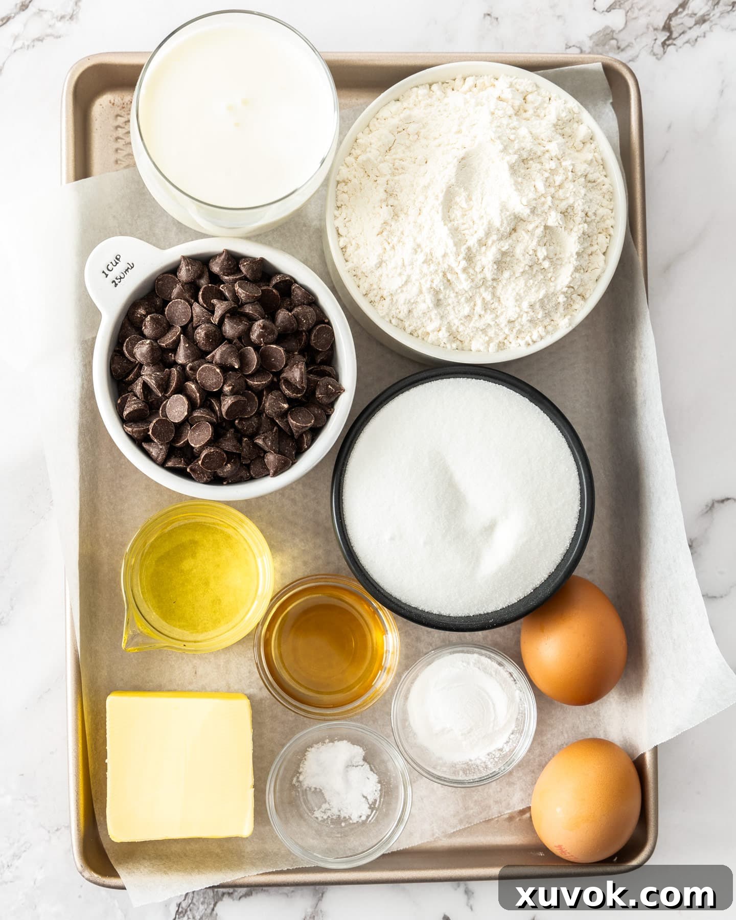 All the necessary ingredients for a homemade chocolate chip loaf cake laid out on a baking tray, including flour, sugar, butter, eggs, buttermilk, and chocolate chips.