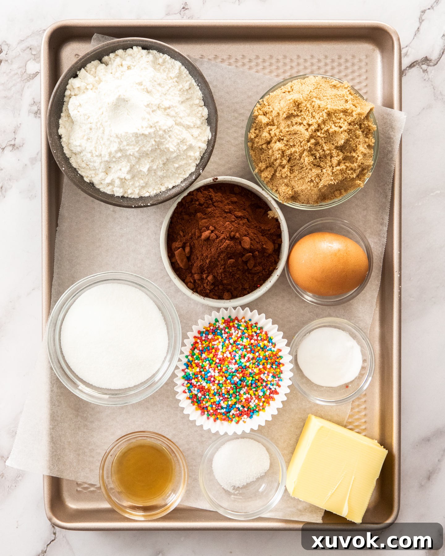 Various ingredients for chocolate sprinkle cookies laid out on a baking tray, including butter, flour, cocoa, sugars, and colorful sprinkles.