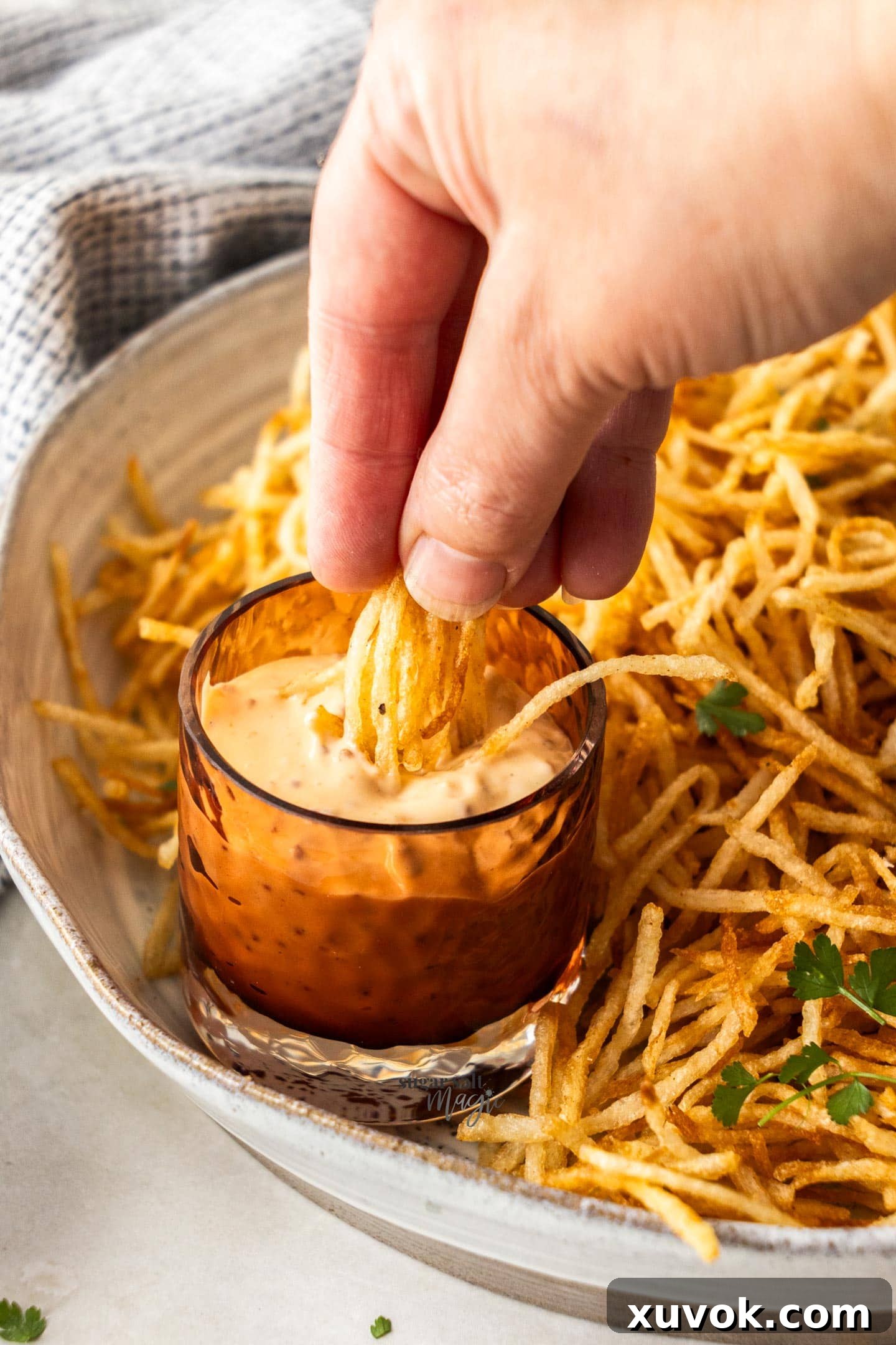 A hand holding a generous handful of crispy shoestring fries, ready to be dunked into a small bowl of dipping sauce. The fries are golden brown and perfectly seasoned.