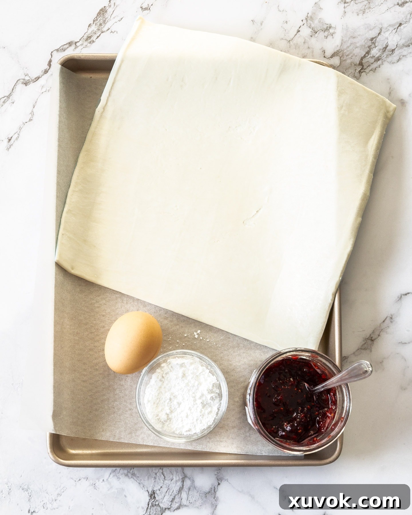 Various ingredients for puff pastry jam tarts laid out: a block of puff pastry, a jar of raspberry jam, an egg, and a bowl of icing sugar.