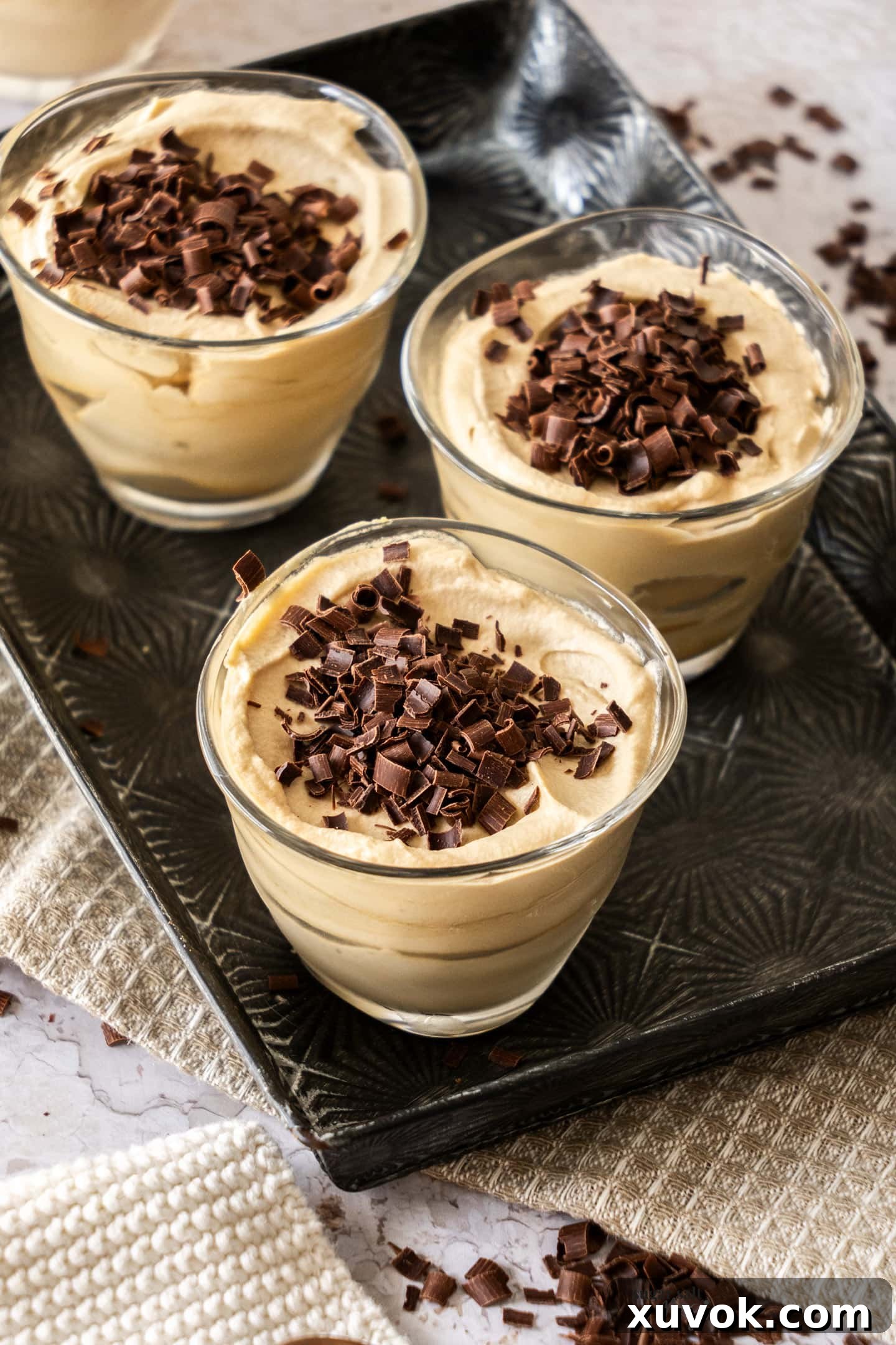 Three decadent coffee mousses beautifully presented in elegant dessert glasses on a rustic metal tray, adorned with chocolate shavings, ready to be enjoyed.