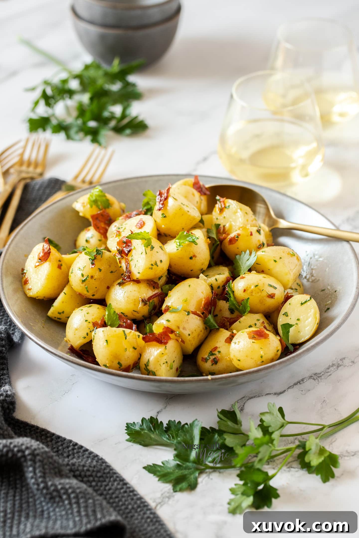 A large grey bowl filled with perfectly cooked baby garlic butter potatoes, adorned with crispy prosciutto and fresh parsley, with gold forks resting nearby, ready to be served.