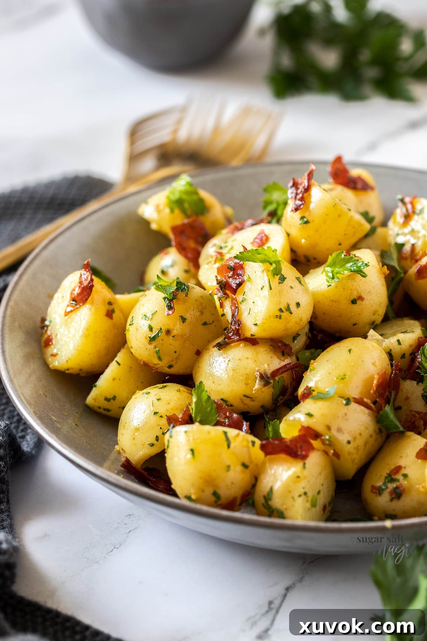 A close-up shot of a grey bowl filled with perfectly cooked baby garlic butter potatoes, generously sprinkled with fresh green parsley and crumbled crispy prosciutto, ready to be enjoyed.