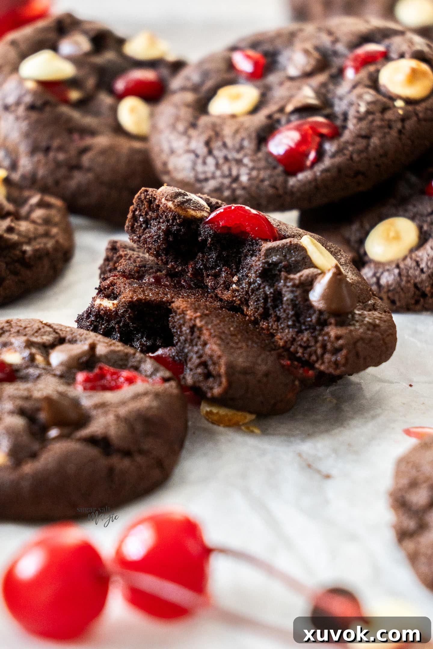 A Black Forest cookie artfully broken in half, revealing its incredibly fudgy, dense chocolate interior dotted with glistening chocolate chips and bright red maraschino cherry pieces.