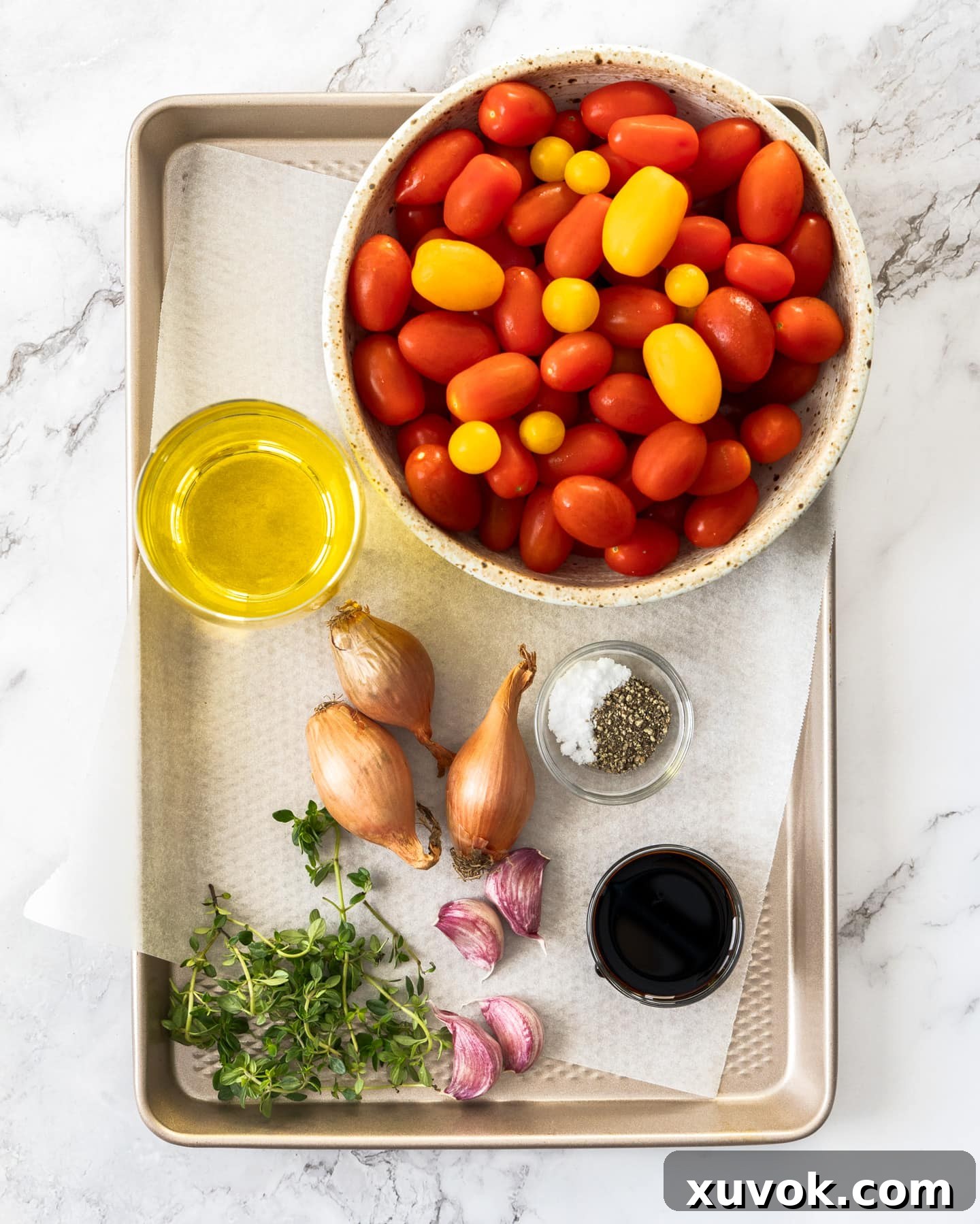 Fresh ingredients for cherry tomato confit, including cherry tomatoes, garlic, shallots, and thyme, arranged on a baking sheet.