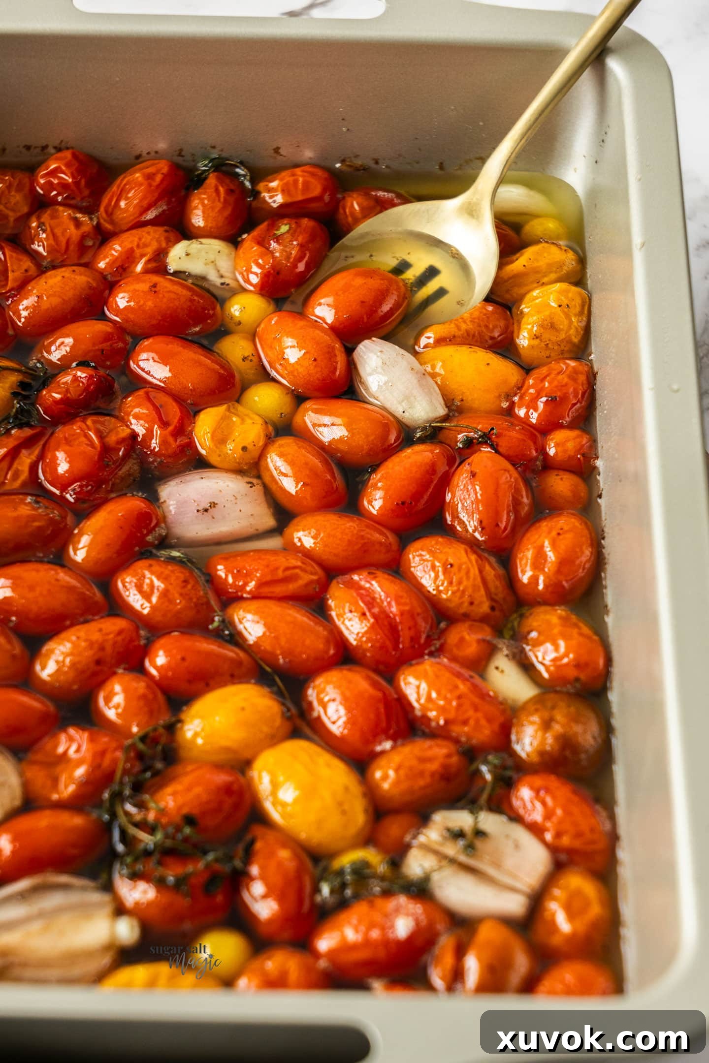 A serving spoon in a rustic pan, scooping up glistening cherry tomato confit with tender garlic cloves and herbs.