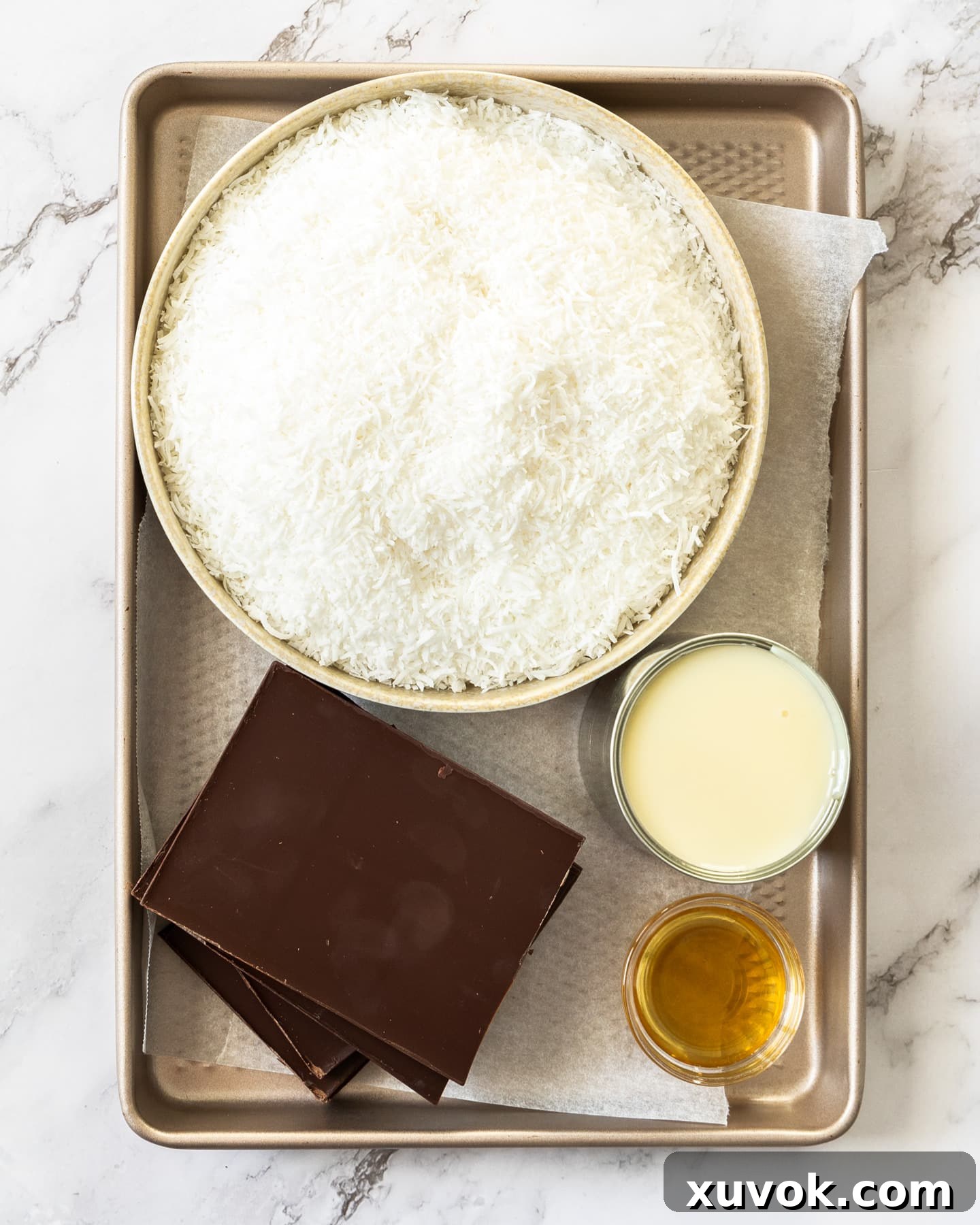A flat lay photograph showcasing the ingredients for coconut balls: shredded coconut, sweetened condensed milk, vanilla extract, and dark chocolate.