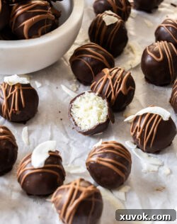A batch of chocolate covered coconut balls with one cut in half showing the inside, on a white plate.