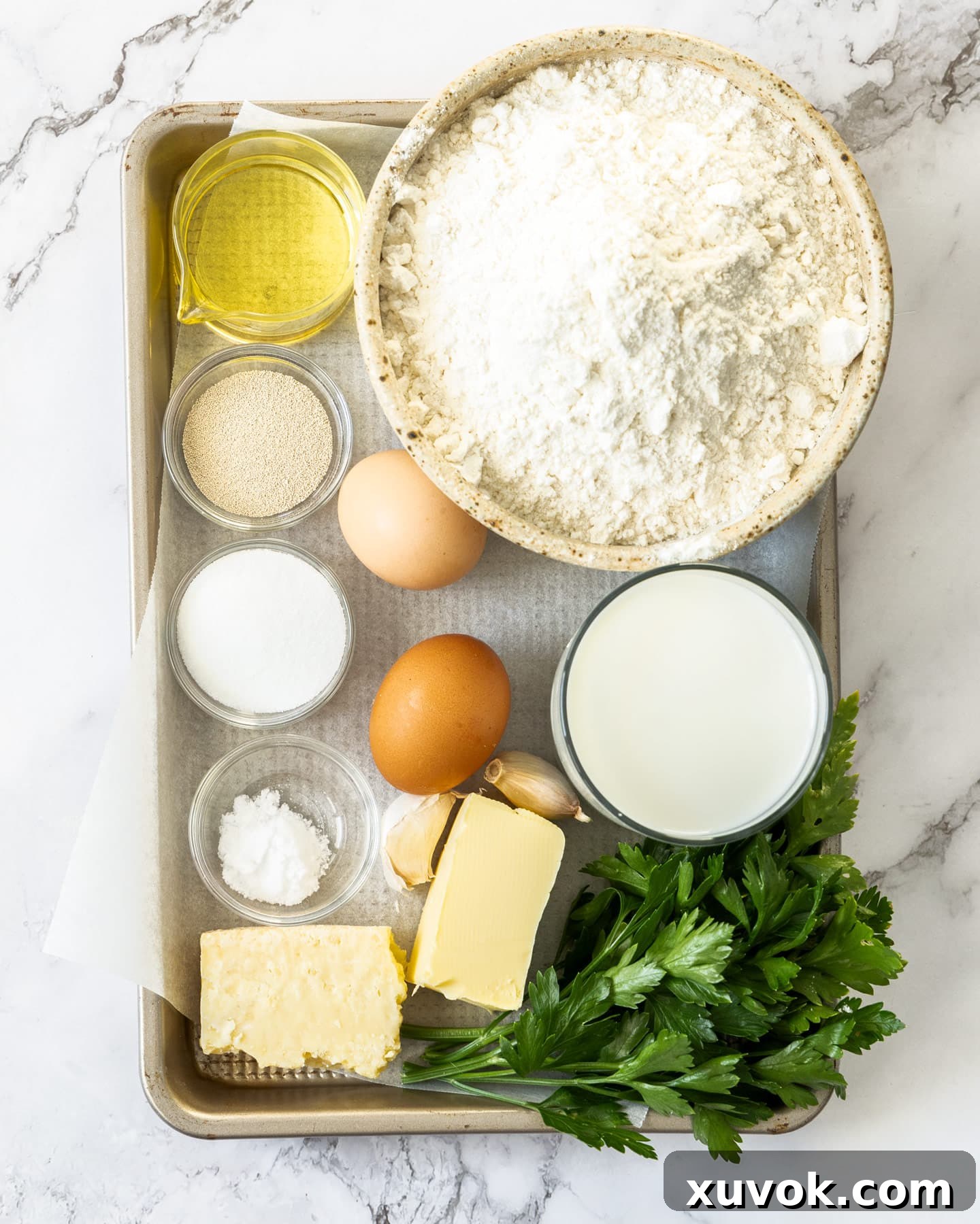 A collection of fresh ingredients for garlic butter dinner rolls, including flour, milk, eggs, butter, garlic, and parsley, neatly arranged on a baking tray.