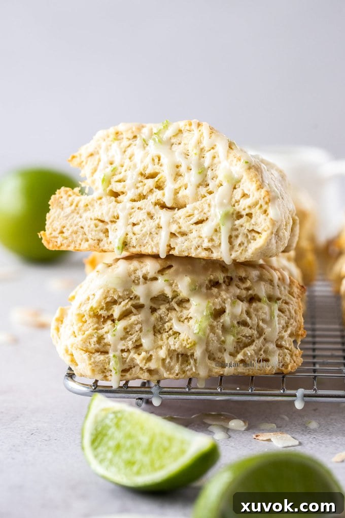 Two perfectly baked lime coconut scones stacked on a wire cooling rack, highlighting their golden edges and crumbly texture. Fresh lime wedges are artfully arranged in the foreground, with a whole lime in the background, all set against a sophisticated grey tabletop, emphasizing the fresh citrus notes of the recipe.