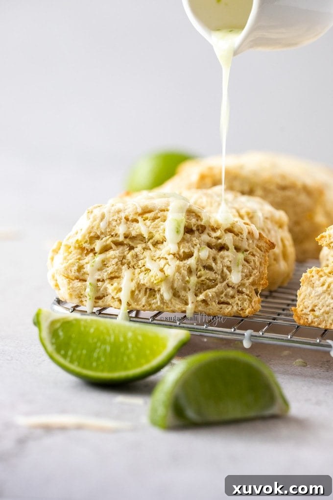A close-up shot of a freshly baked lime coconut scone on a wire rack, with a delicate lime glaze being drizzled over its surface, creating an enticing visual of its crisp exterior and the glistening, tangy topping. The image captures the final, delicious touch of the recipe.