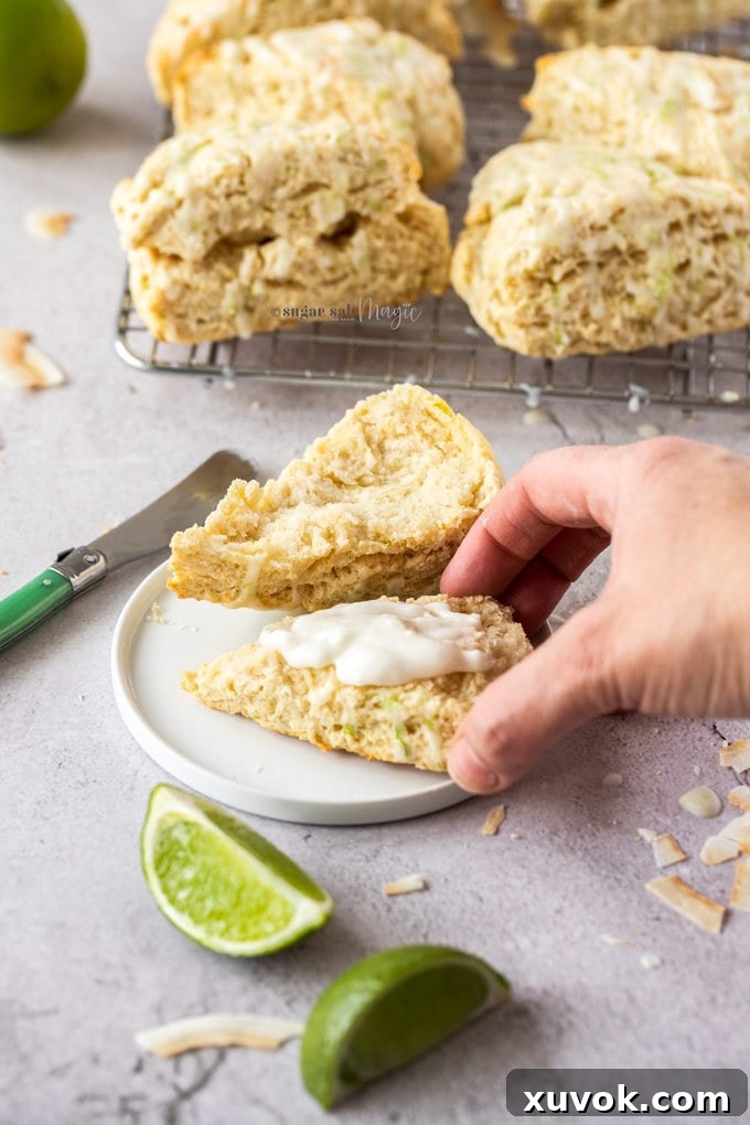 A lime coconut scone split in half, artfully smeared with creamy coconut butter, rests on a pristine white plate. Vibrant lime wedges and delicate coconut flakes are scattered around, with a stack of more scones visible on a wire rack in the background, creating an appealing scene that highlights the delicious serving suggestion for this tropical scone recipe.