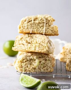 Three perfectly baked lime coconut scones stacked artfully on a wire rack, with vibrant lime wedges nestled in the foreground and a whole lime in the background, all resting on a clean grey bench top. This image showcases the delightful presentation and inviting texture of the tropical scones.