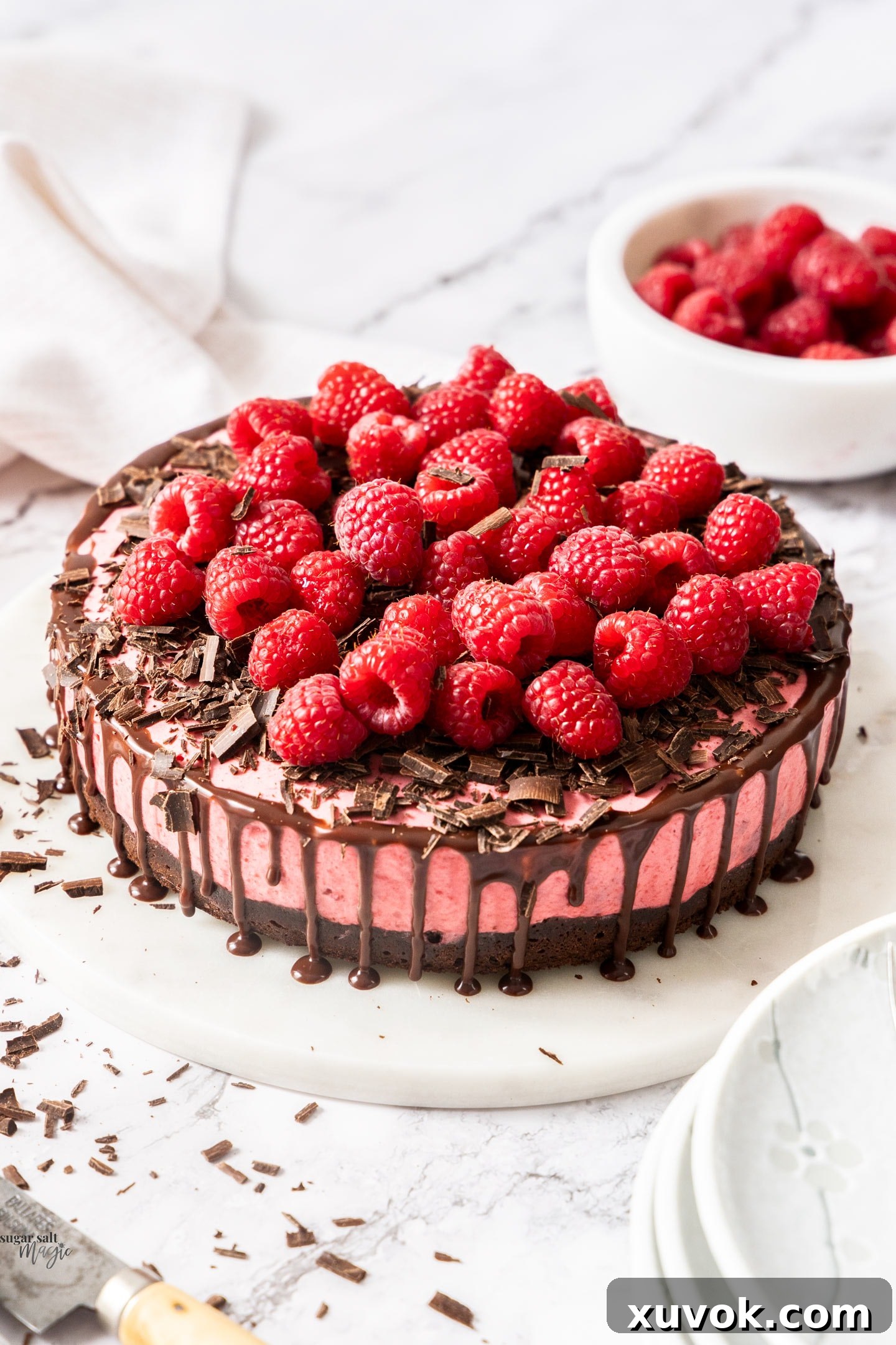 A whole raspberry mousse cake on a cake platter, adorned with fresh raspberries and chocolate shavings.