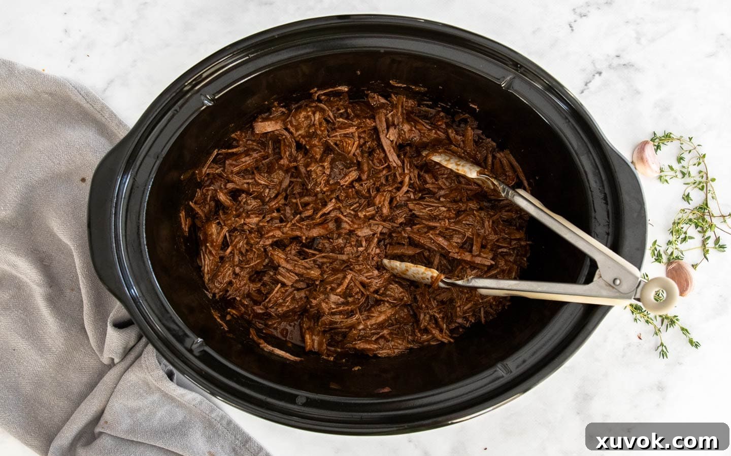 A slow cooker filled halfway with savory shredded beef in gravy, with tongs nearby.