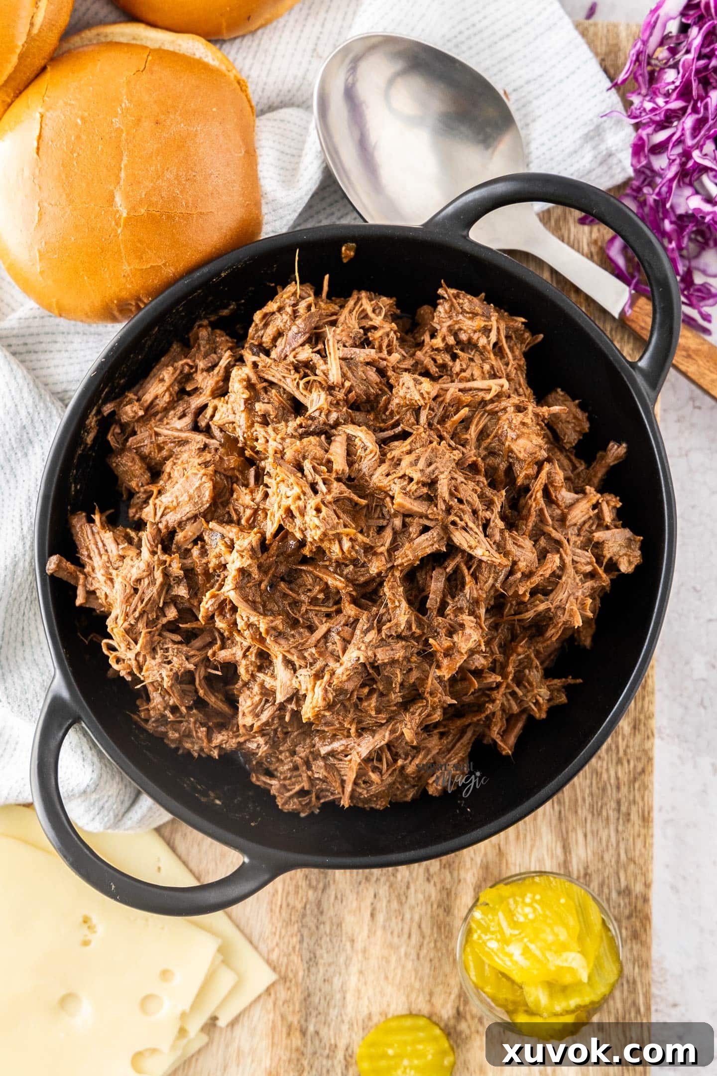 Top-down view of pulled beef in a bowl, surrounded by burger toppings like buns, cheese, and vegetables.