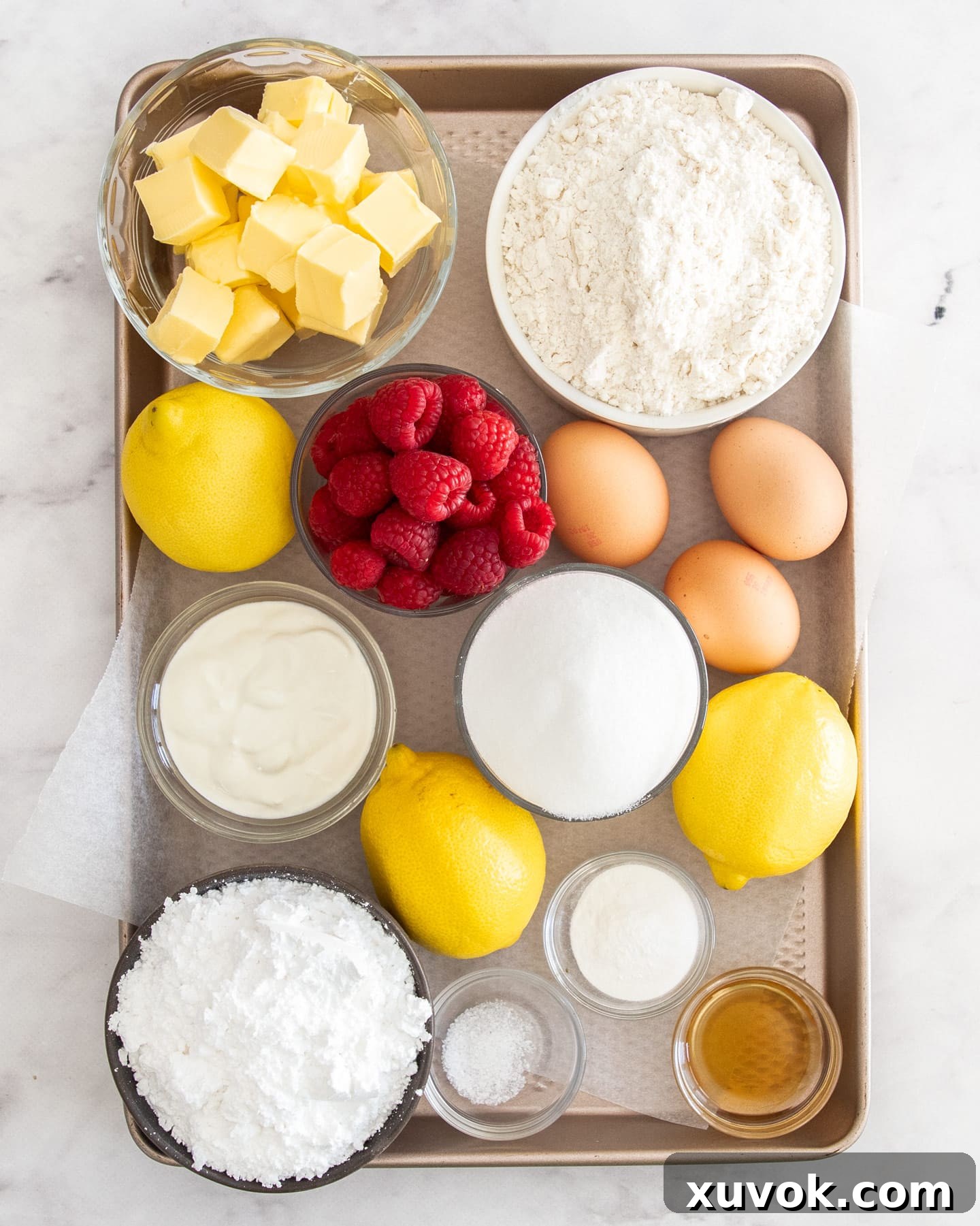 A flat lay photograph showcasing all the fresh and dry ingredients meticulously laid out for baking the raspberry lemon loaf, including flour, sugar, lemons, raspberries, eggs, butter, and Greek yogurt.