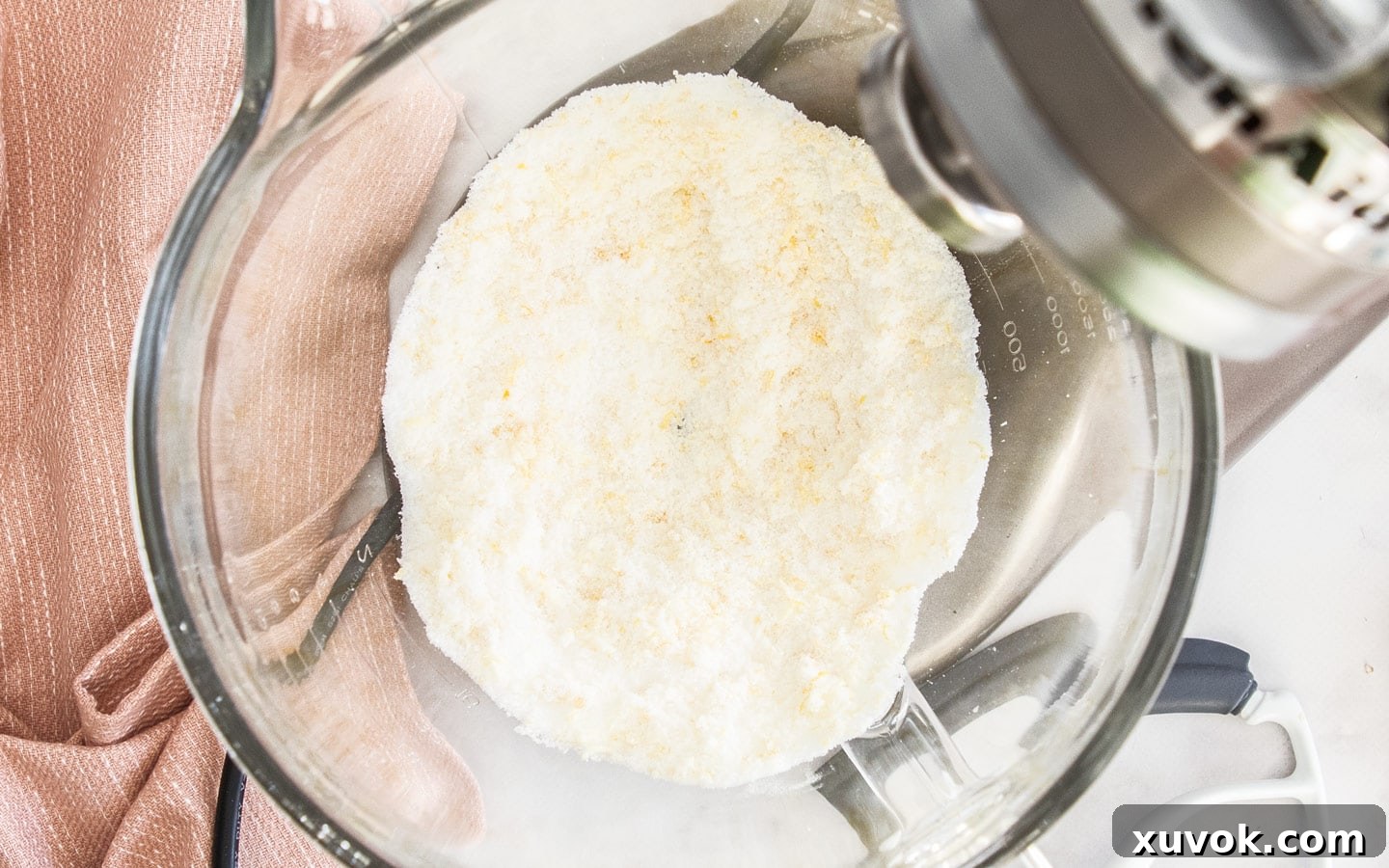 A close-up shot of granulated sugar and bright yellow lemon zest being massaged together in a mixing bowl, illustrating the technique to release lemon oils for intense flavor.
