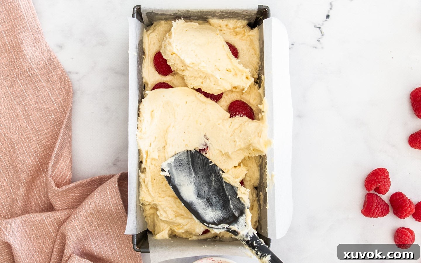 A close-up view of the cake batter and fresh raspberries being carefully layered into a parchment-lined loaf pan before baking, ensuring even distribution.
