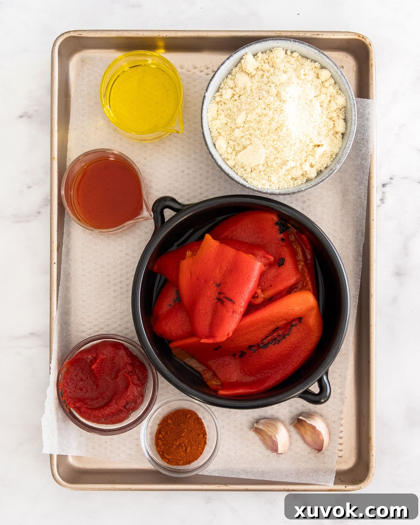 Carefully arranged ingredients for Romesco sauce, including almonds, roasted red peppers, garlic, and a bottle of olive oil, on a rustic baking tray.