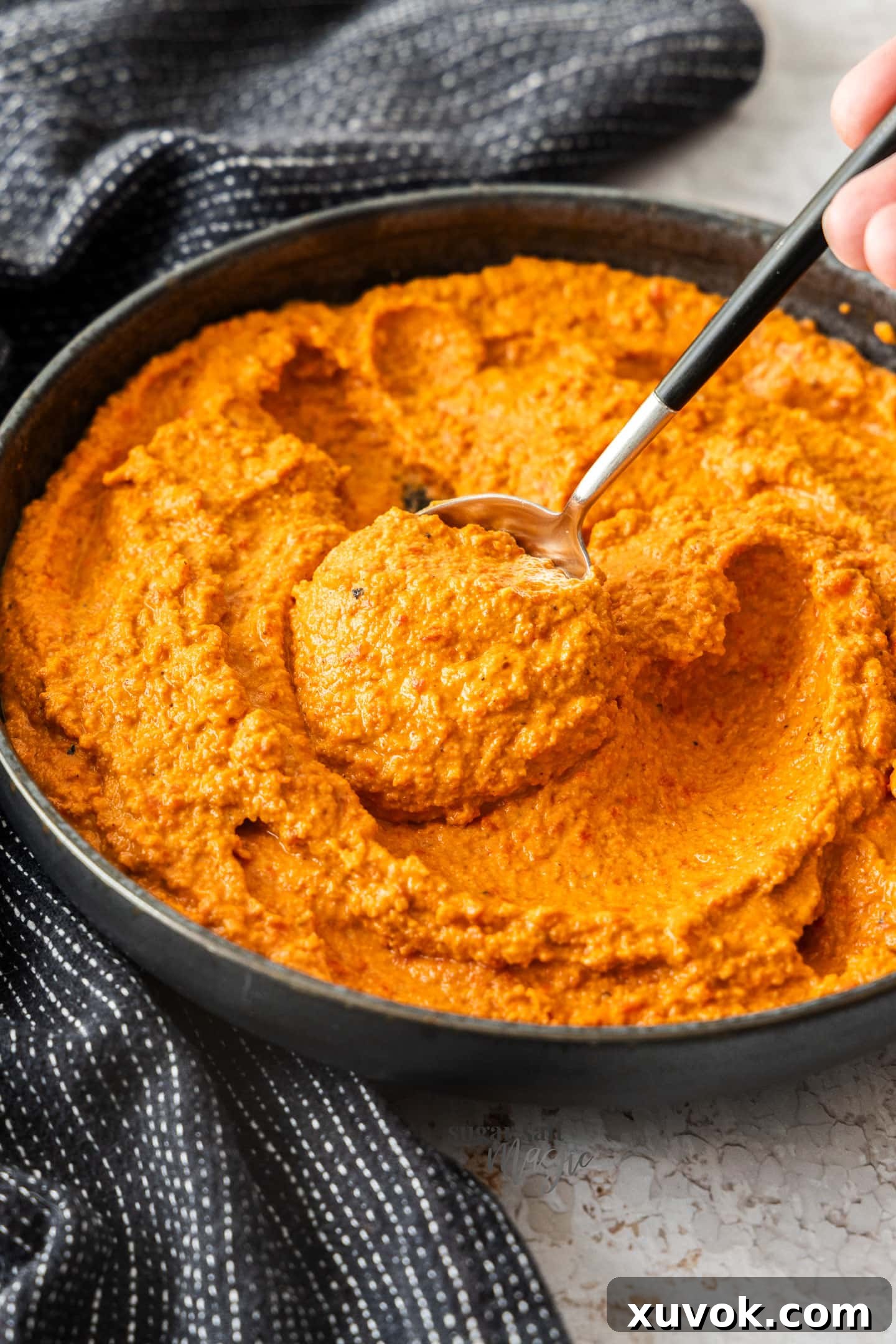 A spoon generously scooping a rich swirl of Romesco sauce from a white ceramic bowl, highlighting its vibrant red color and creamy texture.