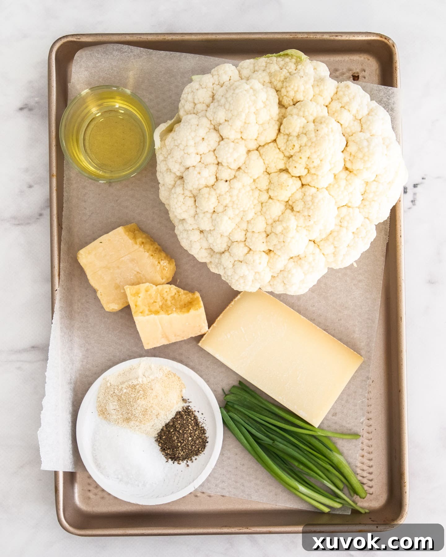 Assortment of ingredients for cheesy cauliflower steaks laid out on a baking tray, including a whole cauliflower, cheeses, oil, and spices.