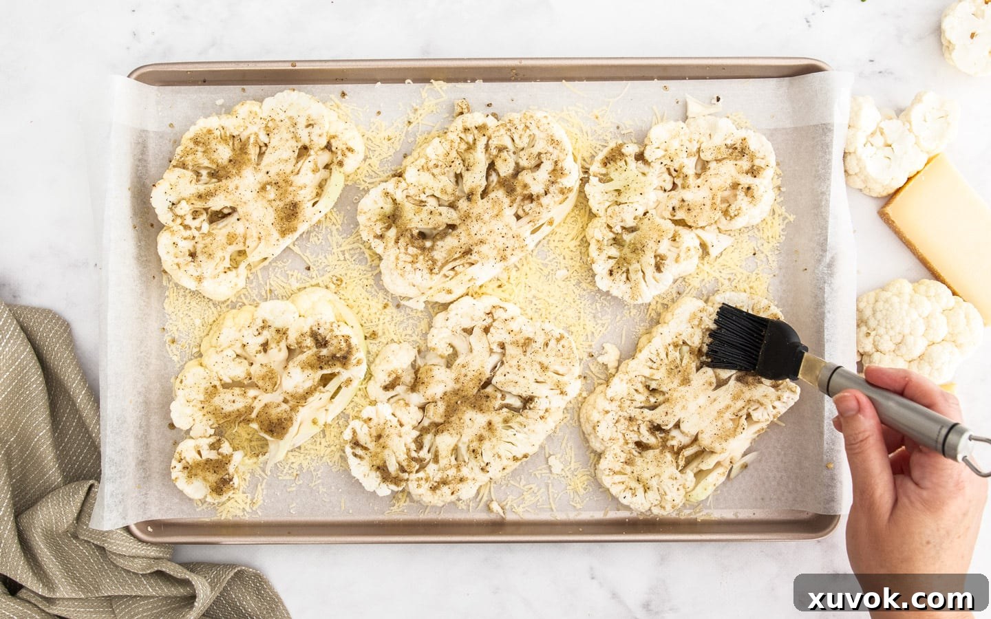 A hand brushing the savory seasoning mixture onto the cauliflower steaks on a baking sheet.