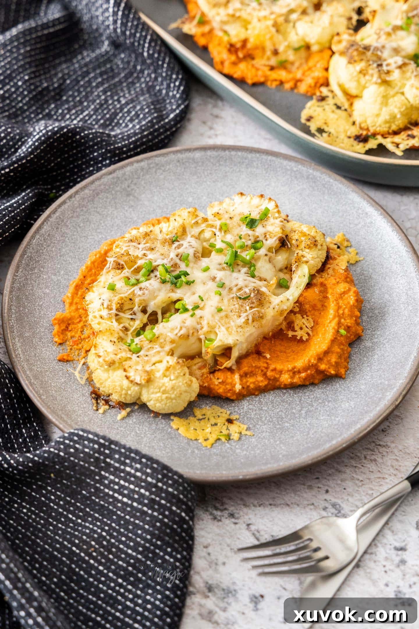 A single cheesy cauliflower steak on a white plate, drizzled with romesco sauce and garnished with fresh chives.
