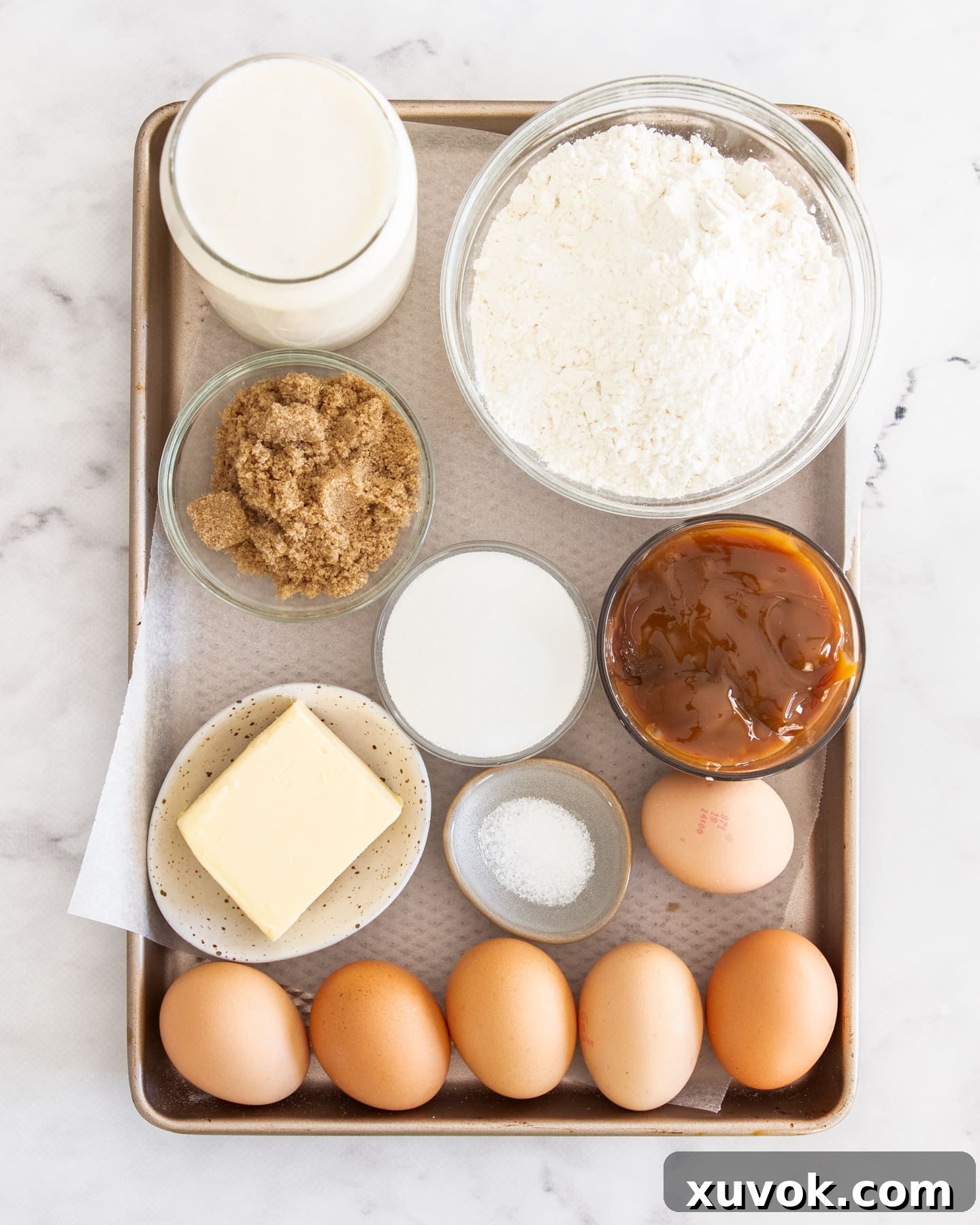 Ingredients for caramel creme brulee tart neatly arranged on a baking tray, highlighting fresh eggs, cream, butter, flour, and a jar of caramel sauce.