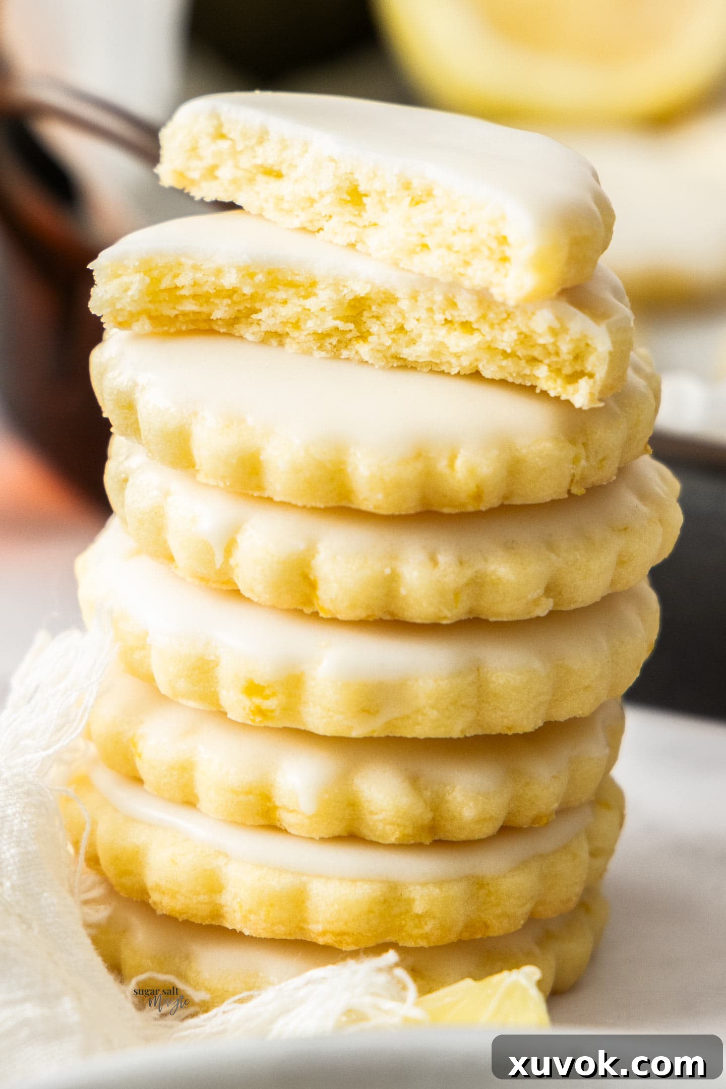 A stack of seven perfectly iced lemon sugar cookies, with the top cookie broken in half to reveal its soft, tender crumb and the vibrant lemon zest flecks within. The glossy lemon glaze adds a tempting sheen.