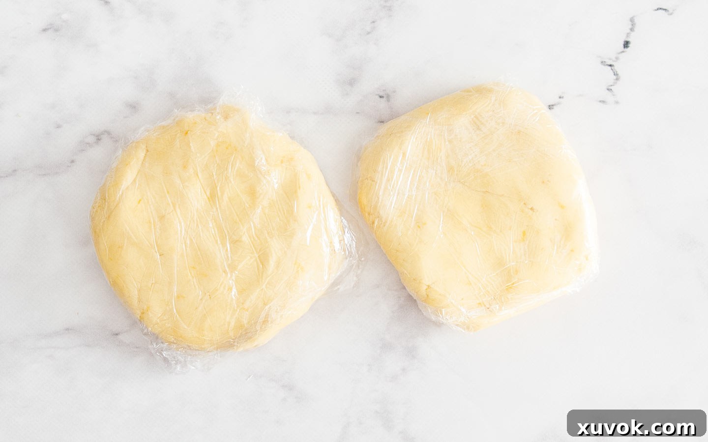Two neatly wrapped disks of lemon cookie dough, securely covered in plastic wrap, ready for chilling in the refrigerator to firm up before rolling and baking.
