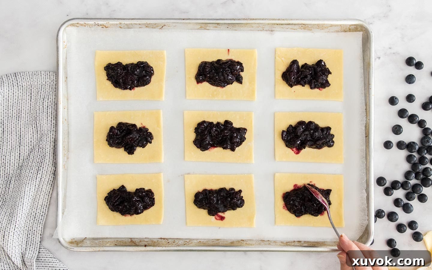 Pastry rectangles on a baking sheet, each topped with a dollop of blueberry jam filling, leaving clean borders.