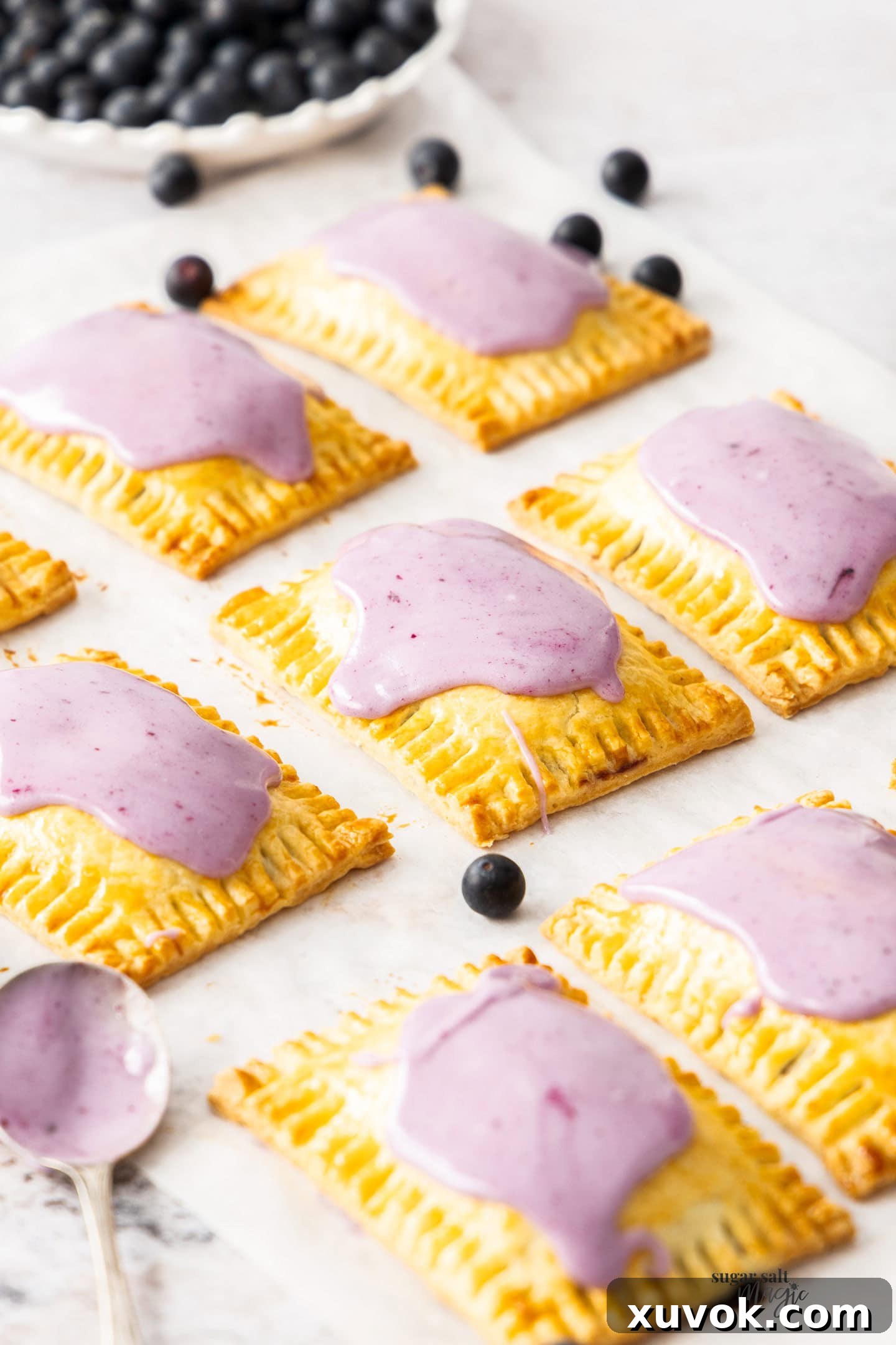 A batch of nine freshly baked and iced blueberry pop tarts arranged neatly on a sheet of baking paper.