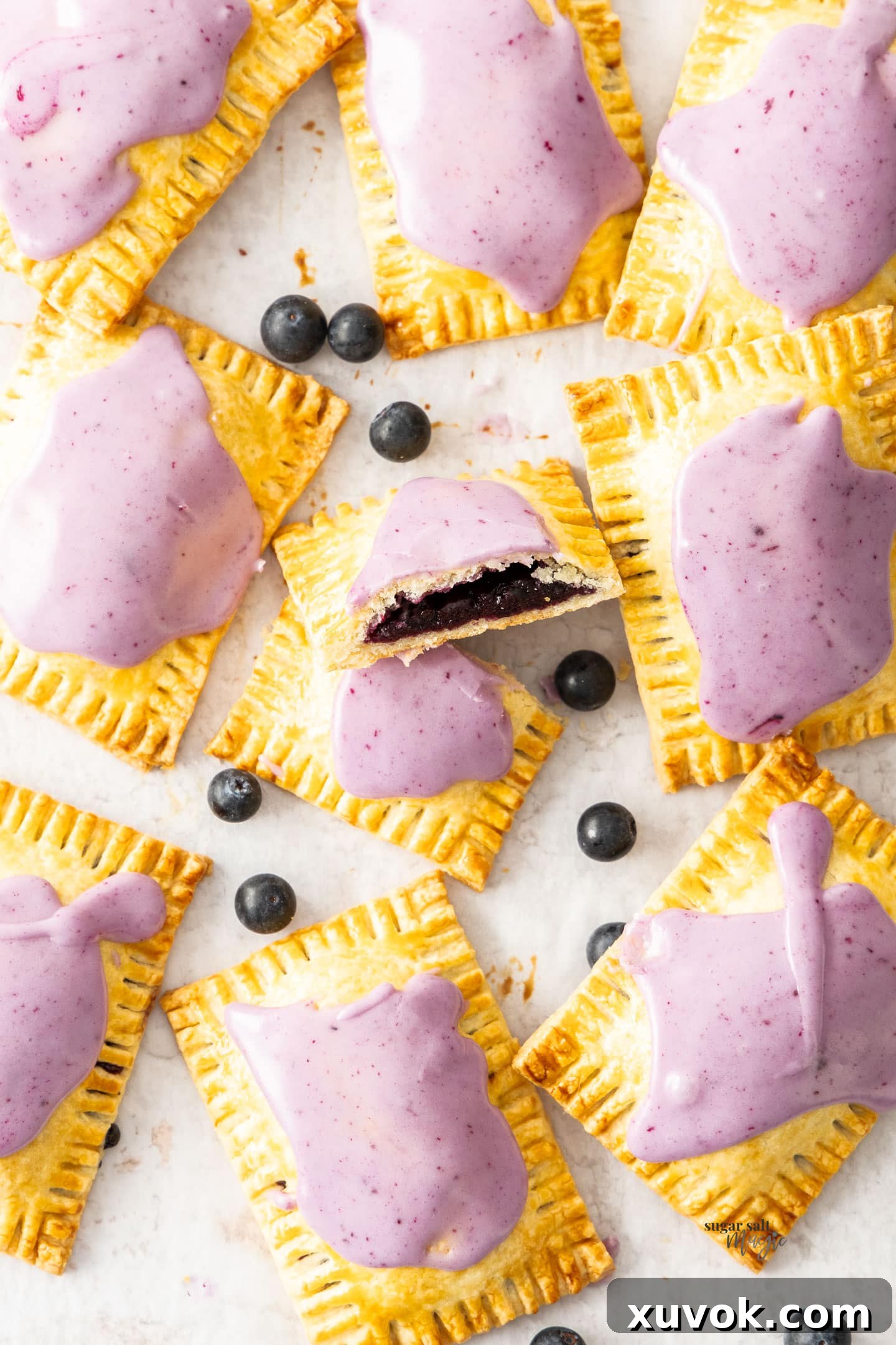A top-down view of a fresh batch of nine homemade blueberry pop tarts on baking paper, with one cut in half to show the filling.