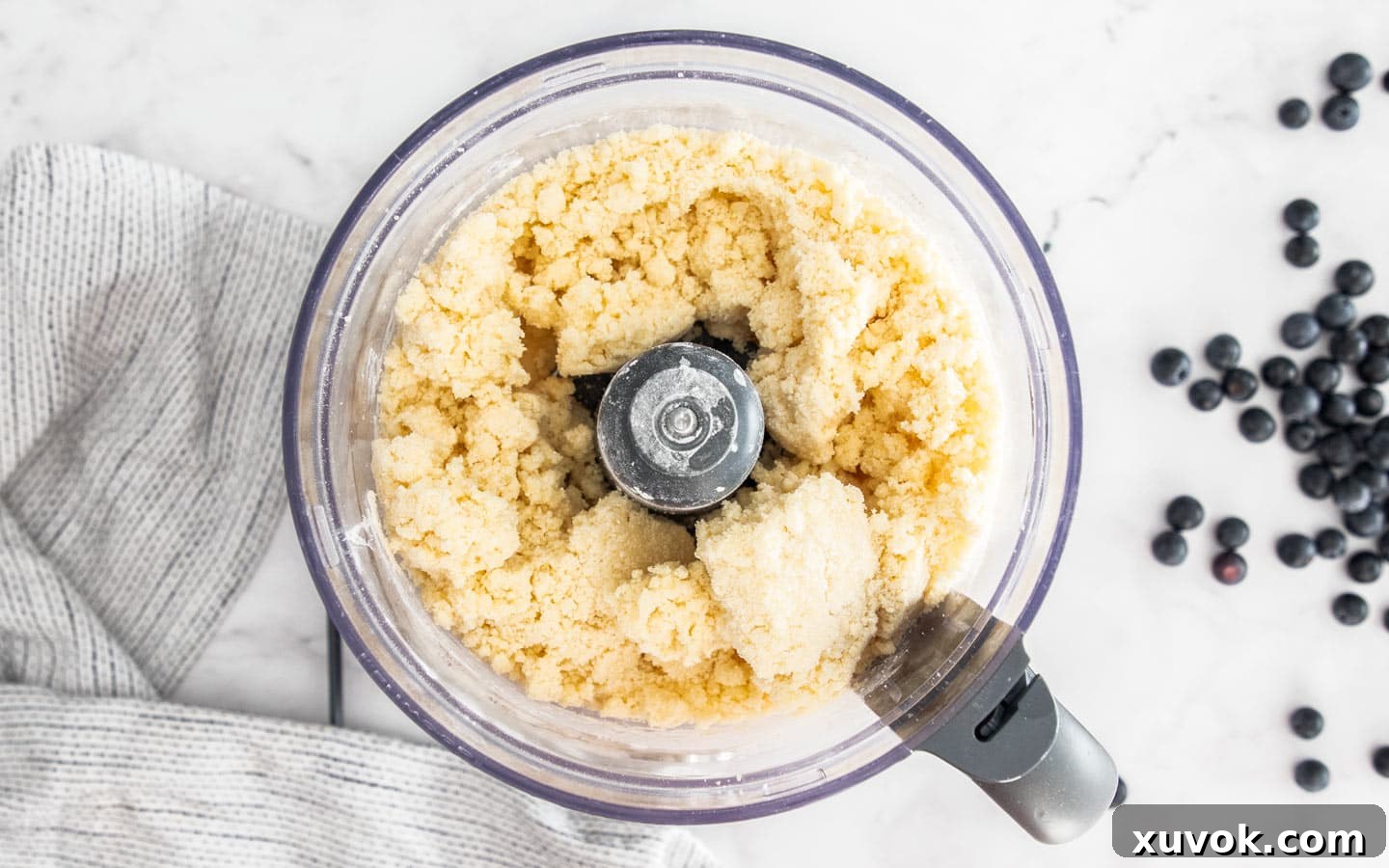 The pastry mixture forming large clumps in a food processor bowl after adding cold water, indicating the right consistency.