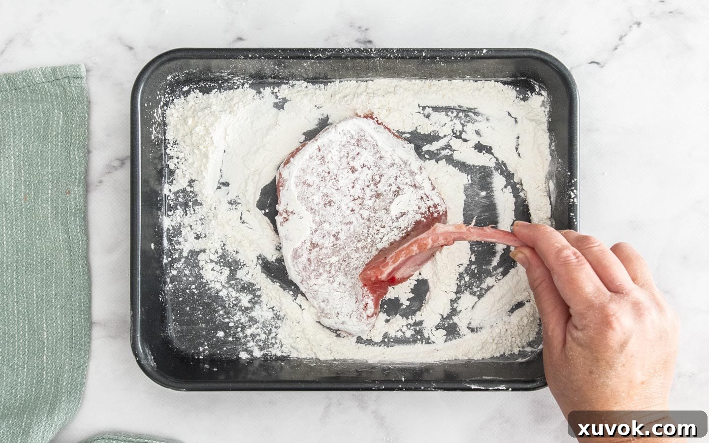 A raw pork cutlet being coated in a shallow bowl of flour, demonstrating the first step of the breading process.