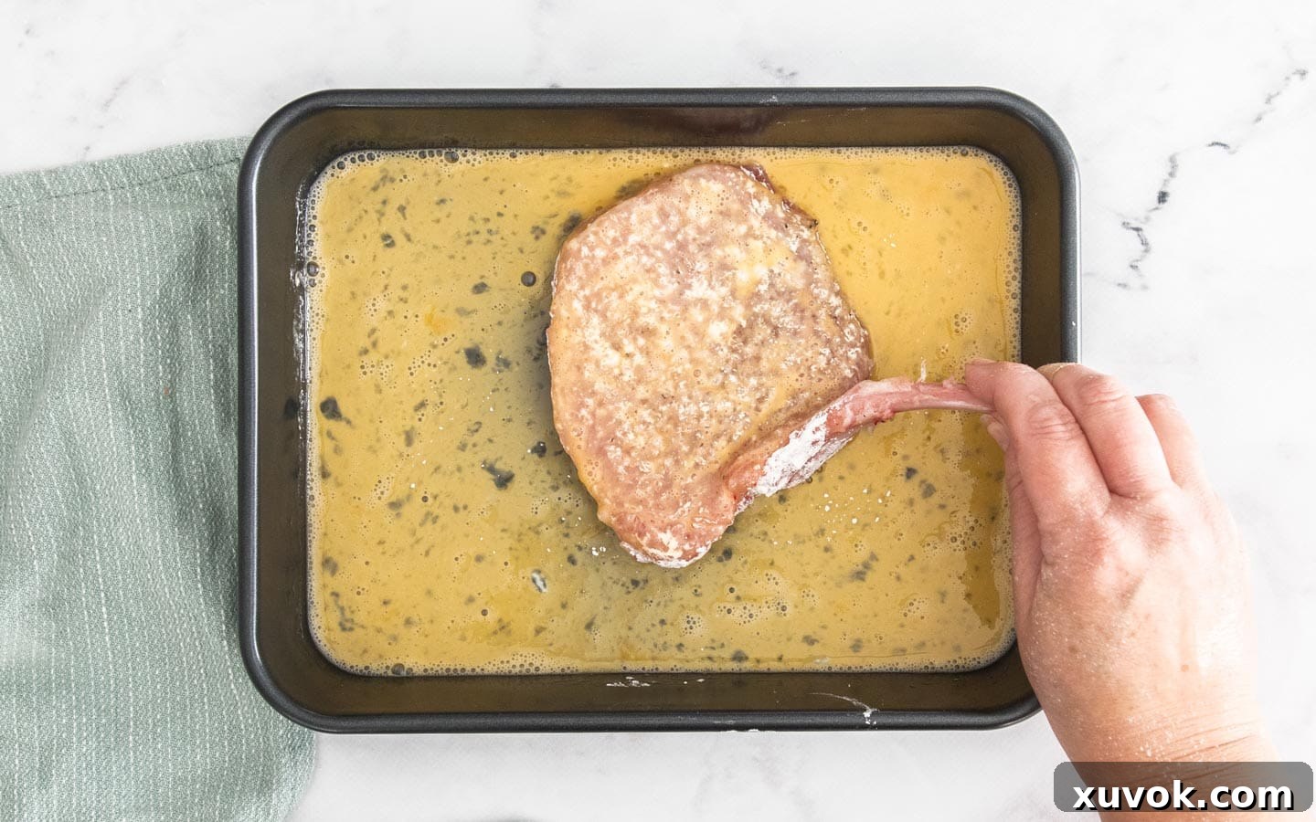 A floured pork cutlet being dipped into a bowl of whisked eggs, ensuring full coverage.